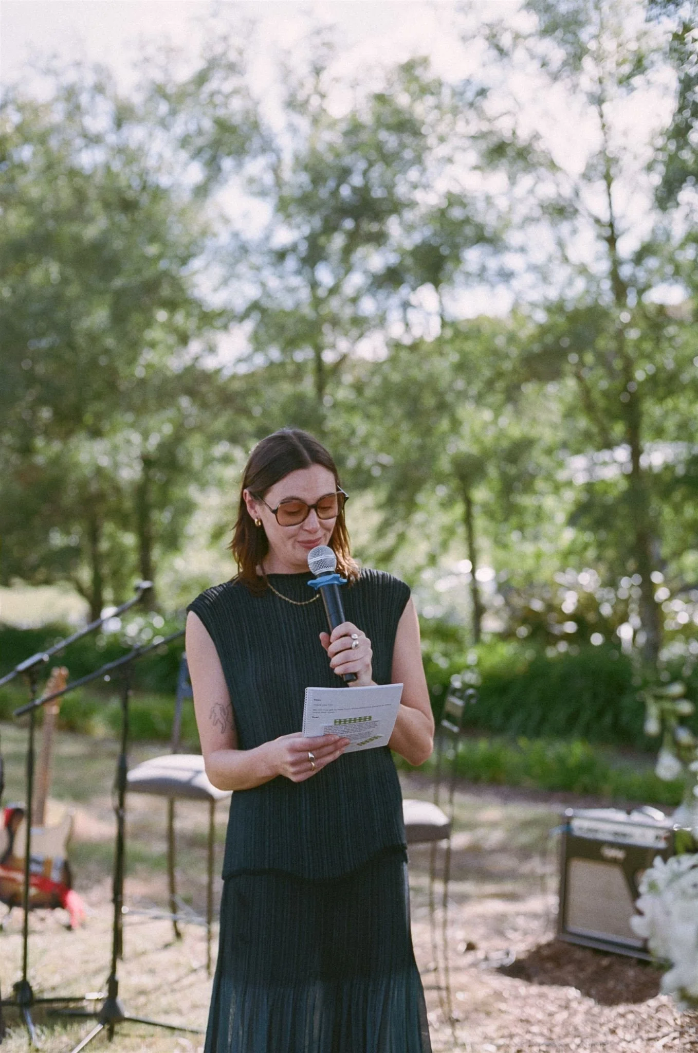 A friend is reading a poem at a wedding ceremony. She wears a delicate black dress and sunglasses.