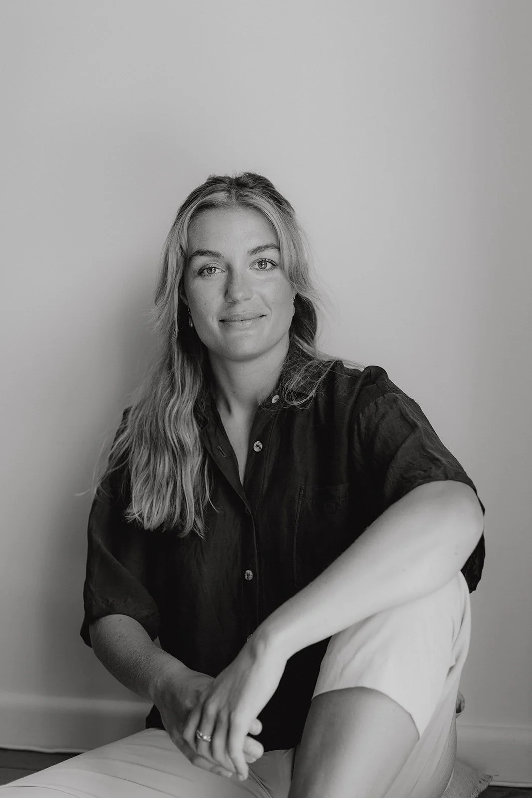 a black and white headshot of a lady sitting with one leg crossed against a wall. She is wearing a dark blouse and smiling.