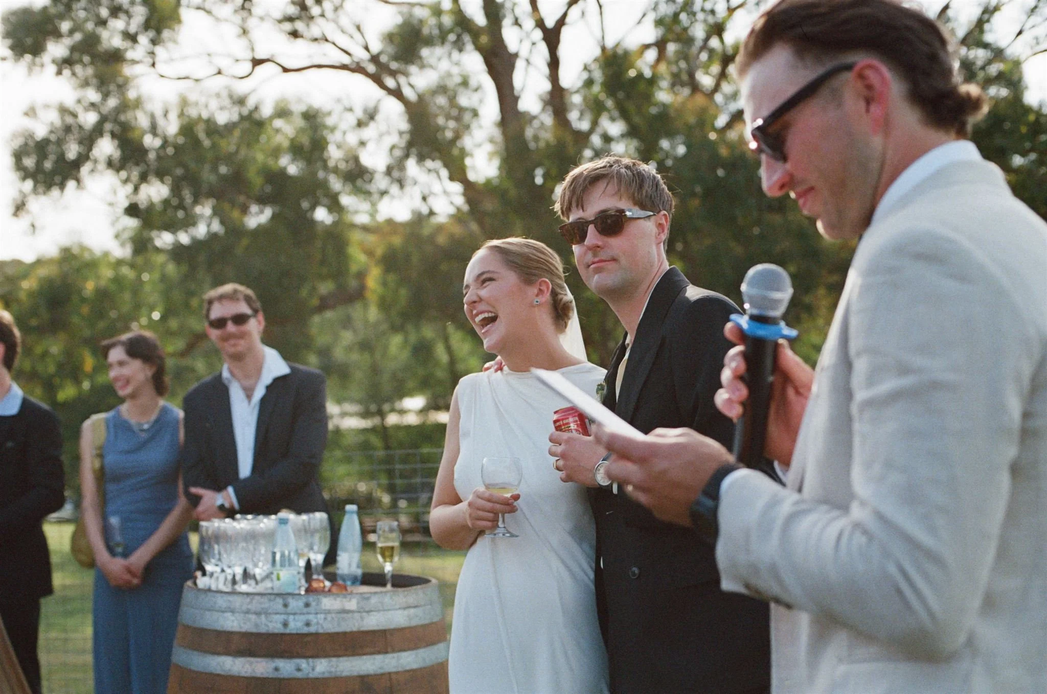 A guest is giving a speech at a wedding while the bride and groom stand in the background smiling. The bride is laughing. 