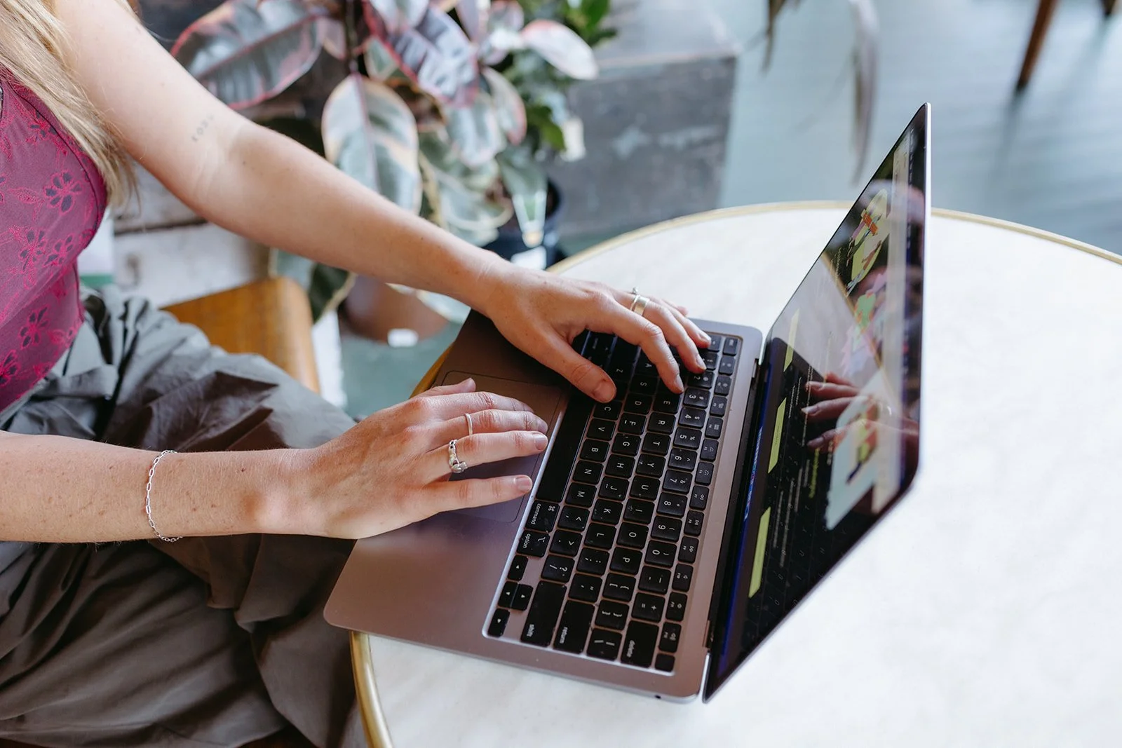 a women's hands type on a laptop, she is wearing a plum top and green trousers.