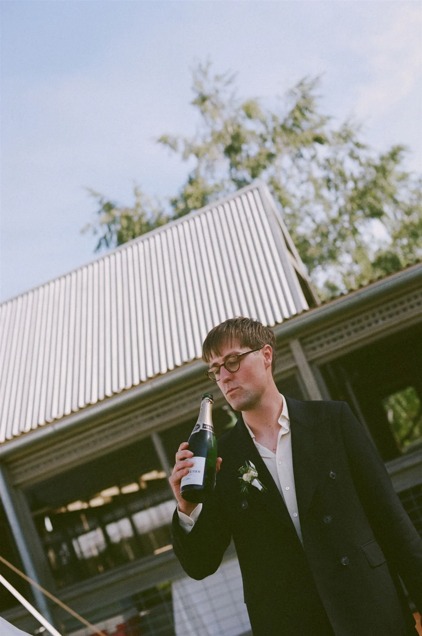 A groom is about to take a swig of champagne straight from the bottle. The sky is blue behind him and he is wearing a black suit. 