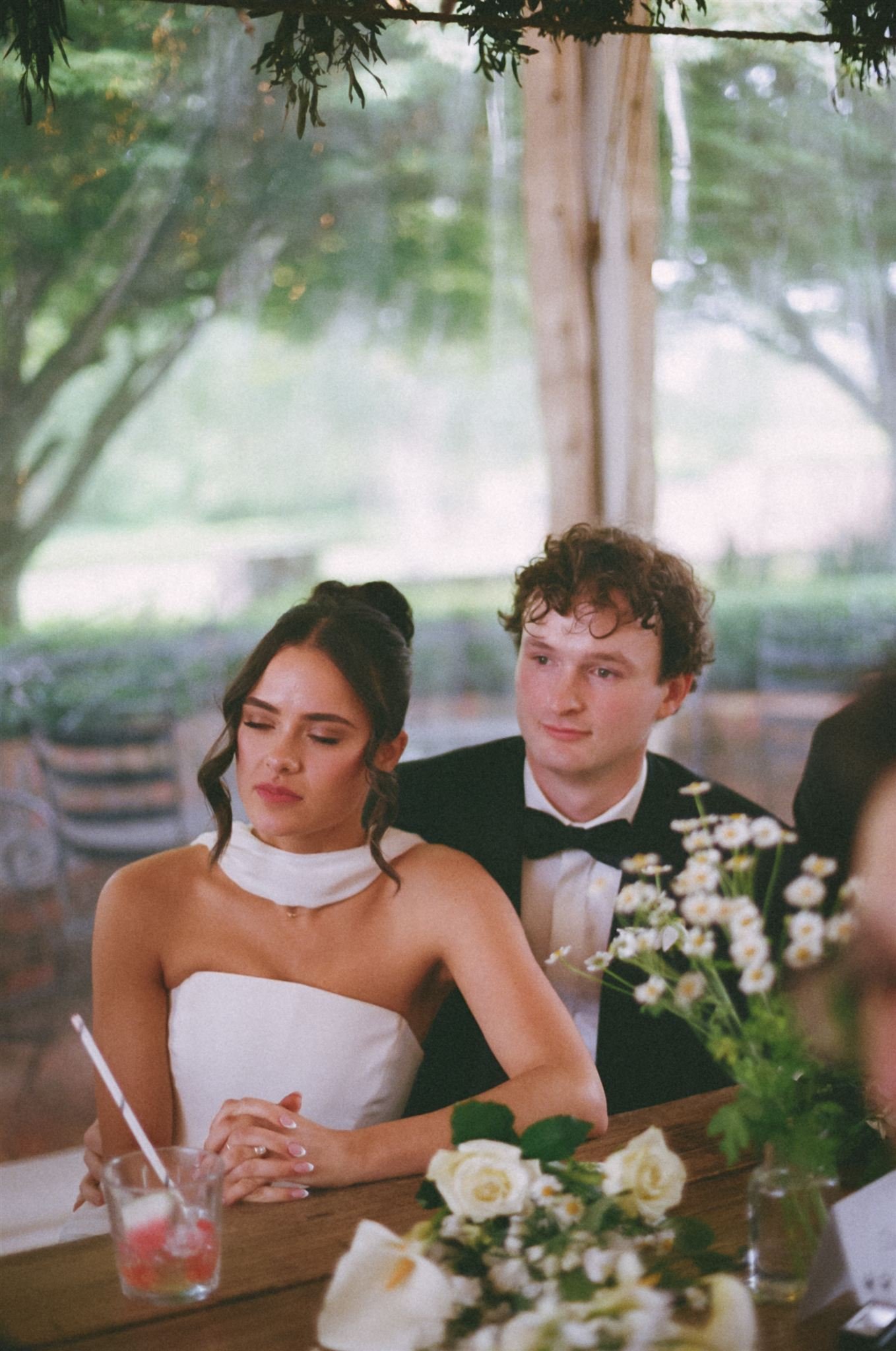 A bride and groom sitting at a table during a wedding reception, with flowers and a drink in front of them. The couple looks emotional while listening to speeches. The background features large windows and natural greenery.
