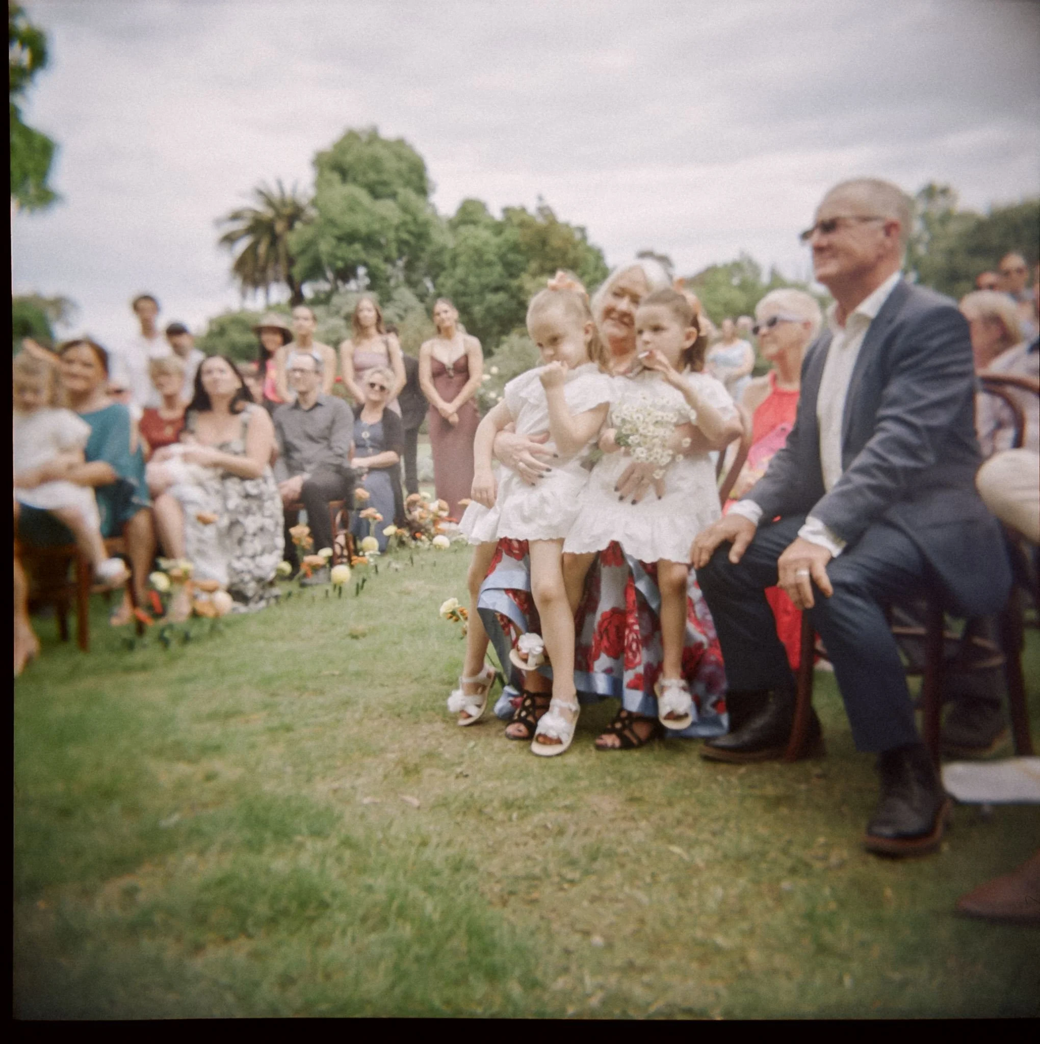 a photograph of a wedding ceremony. The guests are sitting in the park and the two flowers girls sit on their grandfathers lap listening.