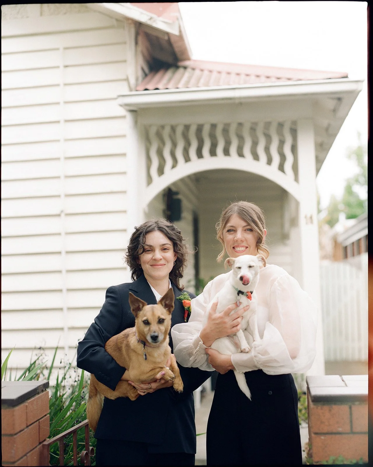 two brides stand outside of their house holding their two small dogs, one bride is in a white blouse and the other is in a dark suit.