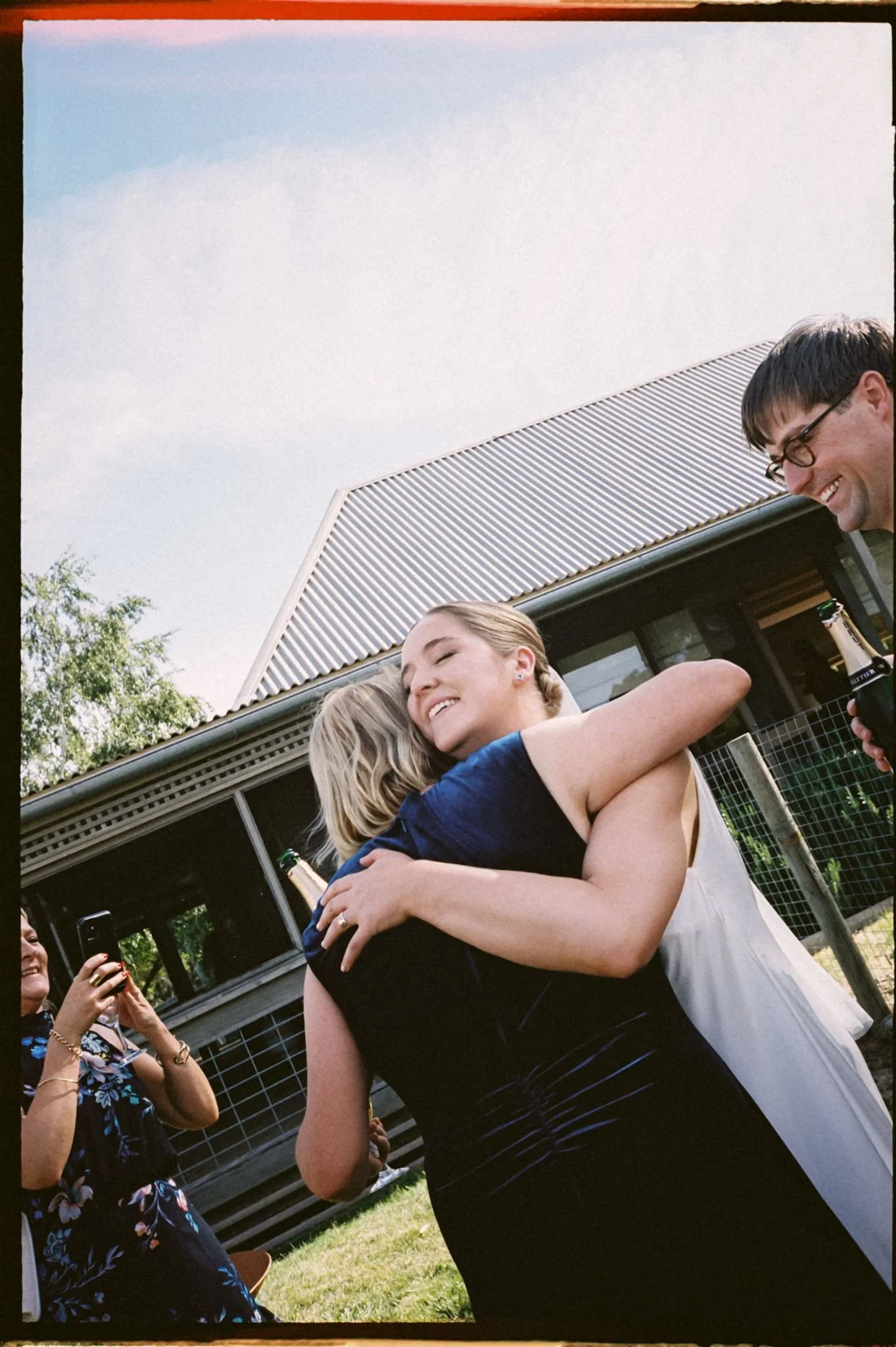 Two women hugging at a wedding, with a man smiling and holding a drink nearby, and another woman taking a photo, in front of a house with a metal roof