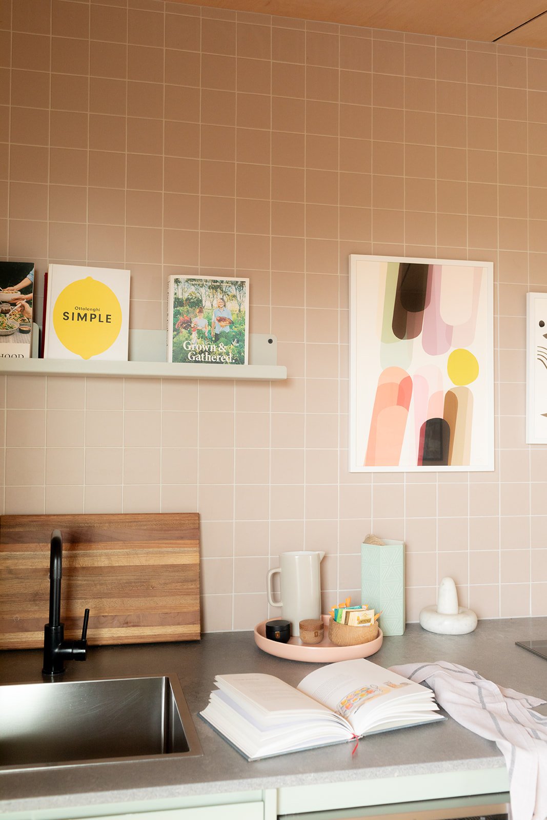 a kitchen bench top with peach tiled splash back, there is art on the walls and a book on the bench with a tea towel.