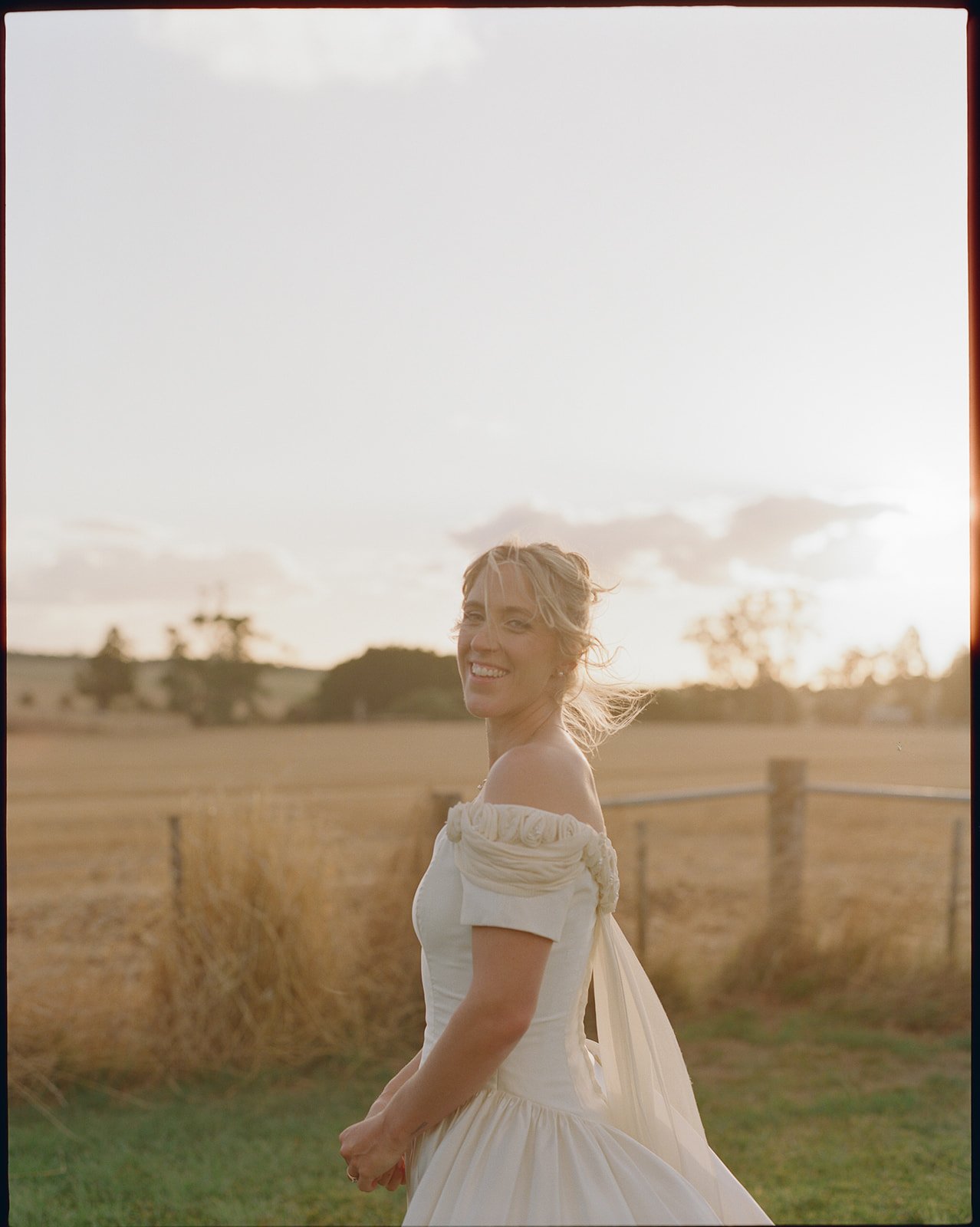 A bride stands with a field in the background, the sun is setting behind her with a warm glow. The wind is blowing her hair. 
