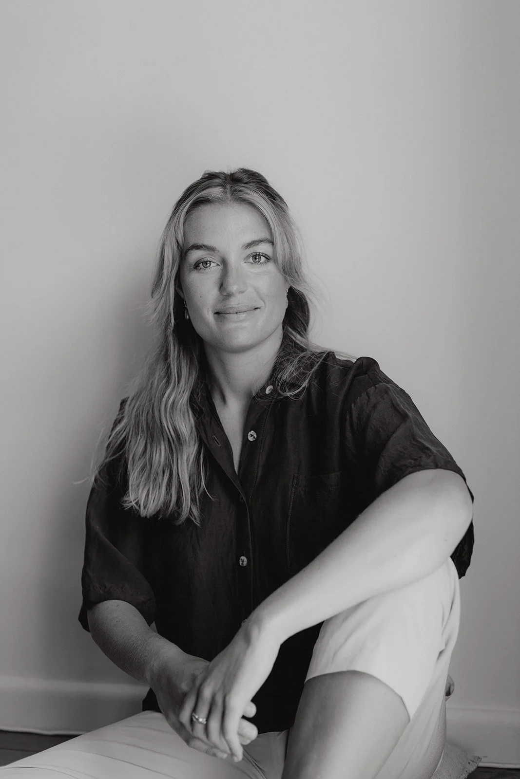 a women sits against a wall, with her arm resting on a bent knee, she wears a dark shirt and light linen pants. she has a soft smile.