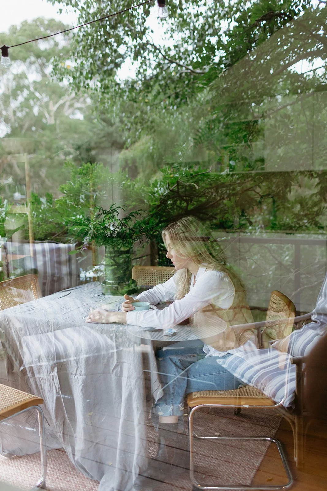 a women sits at a table holding a cup of tea, there is a vase on the table with greenery in, the photo of the women is taken through a window.
