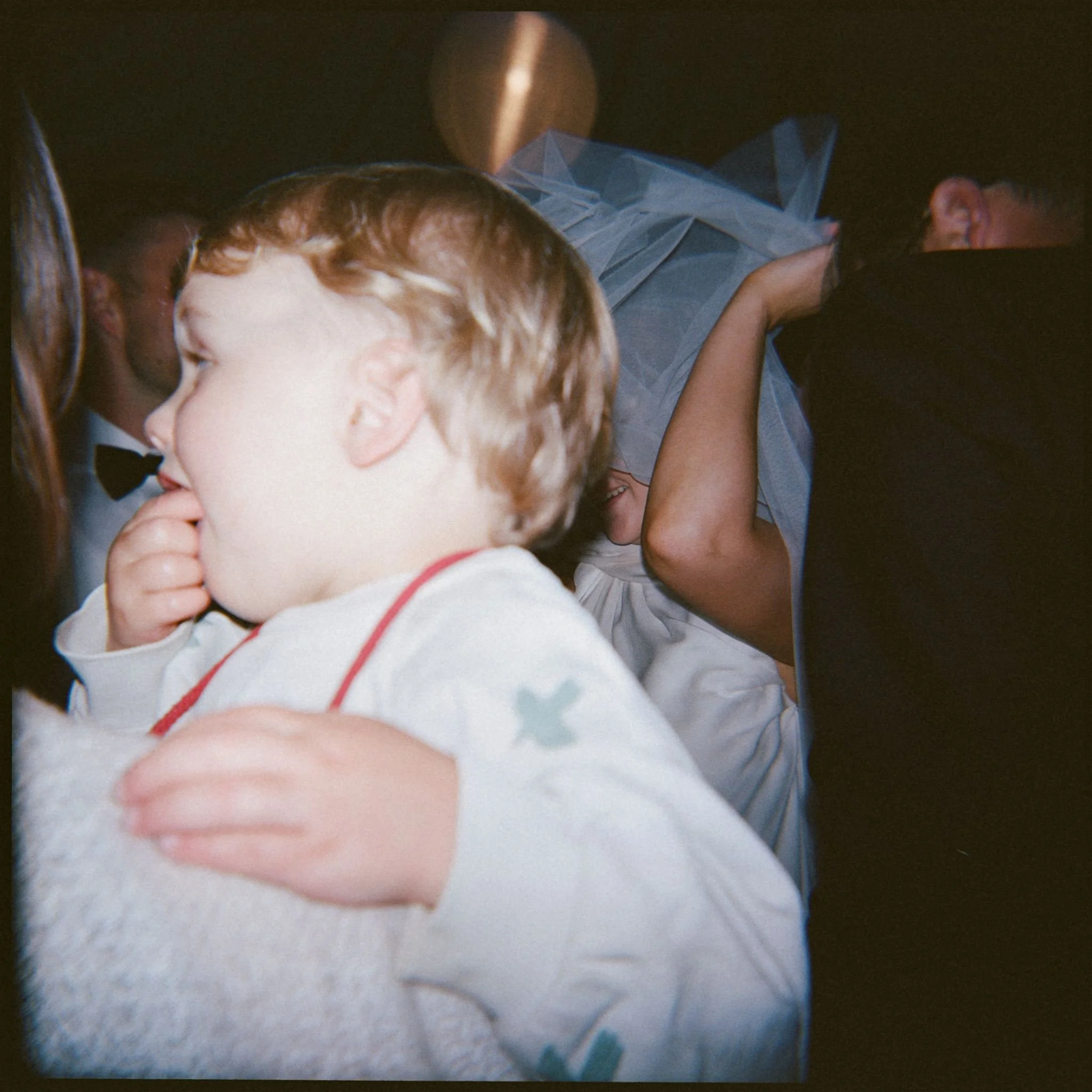 A wedding guest holds a toddler in her arms on the dance floor, the bride is adjusting her veil in the background. 
