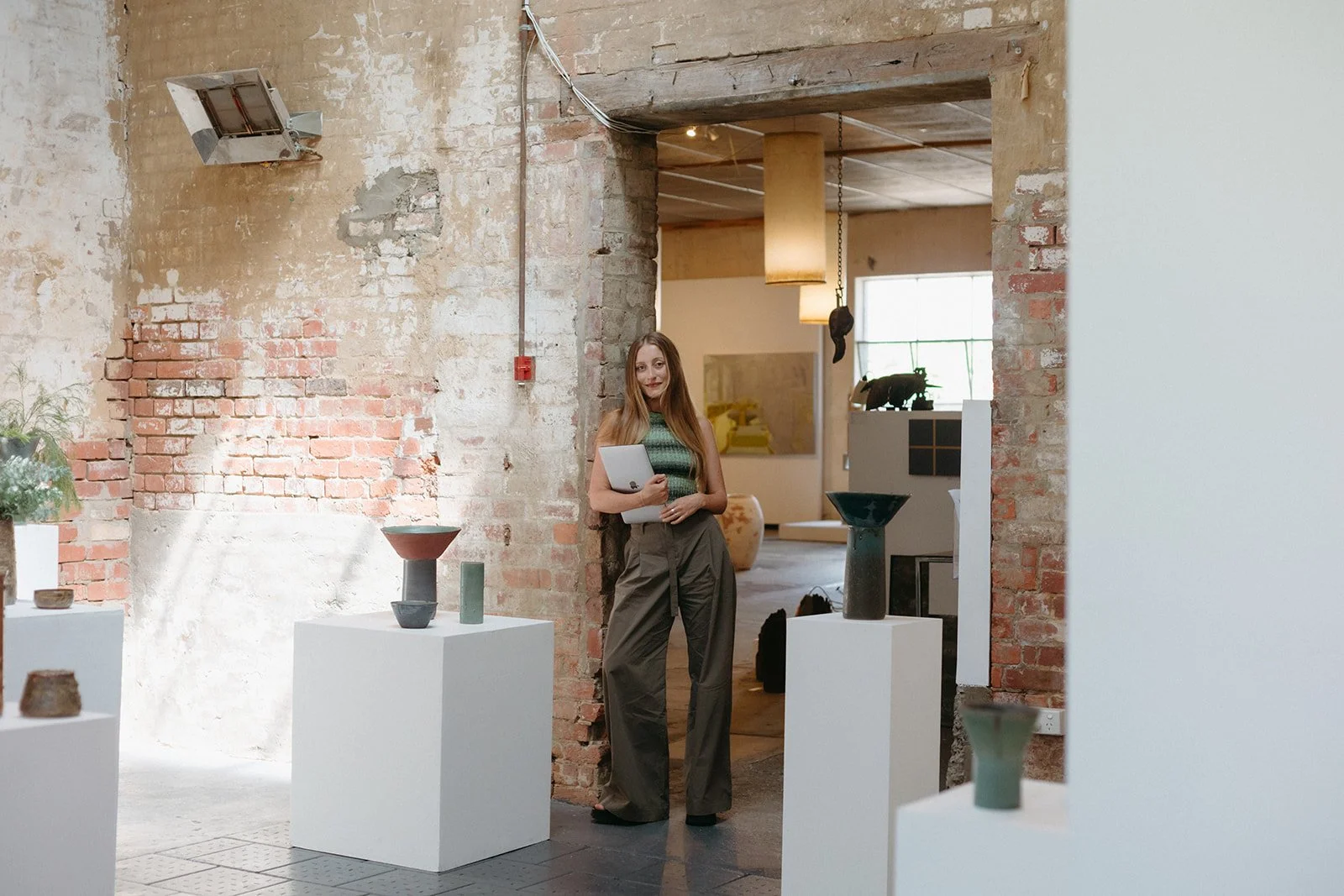 A women stands in an art gallery in Kyneton, she is holding a laptop while the sun shines down through a skylight.