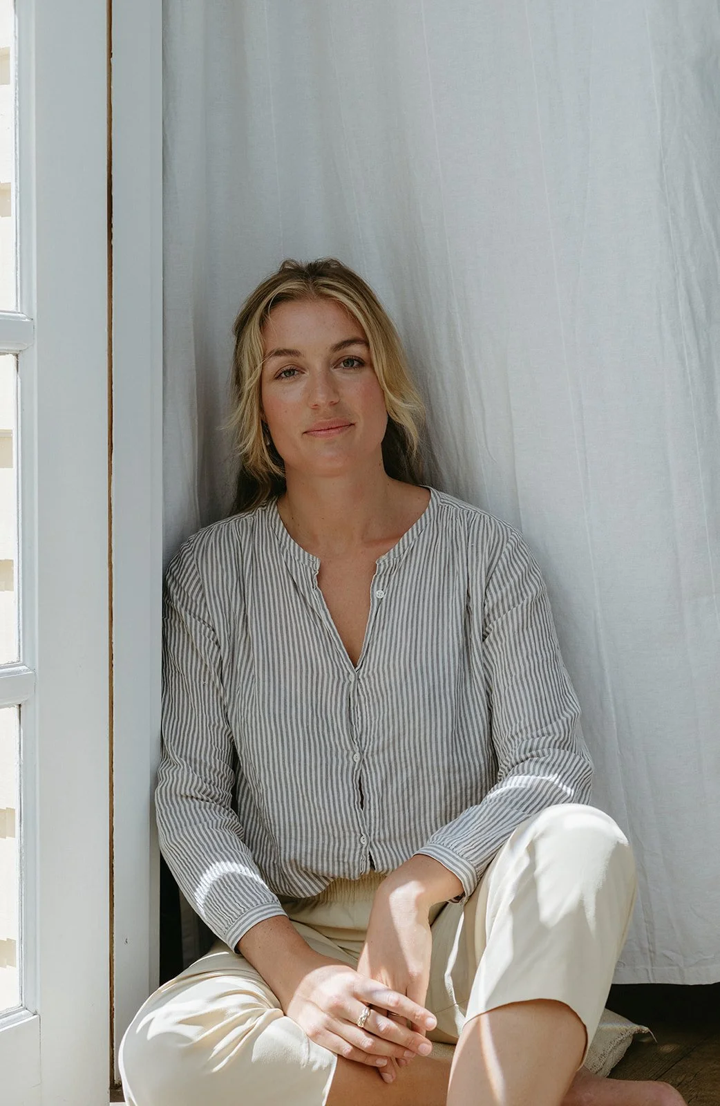 a women sits crossed legged on a step, a linen curtain blows behind her. She is wearing a striped linen shirt and light pants. She has blonde hair and a soft smile.