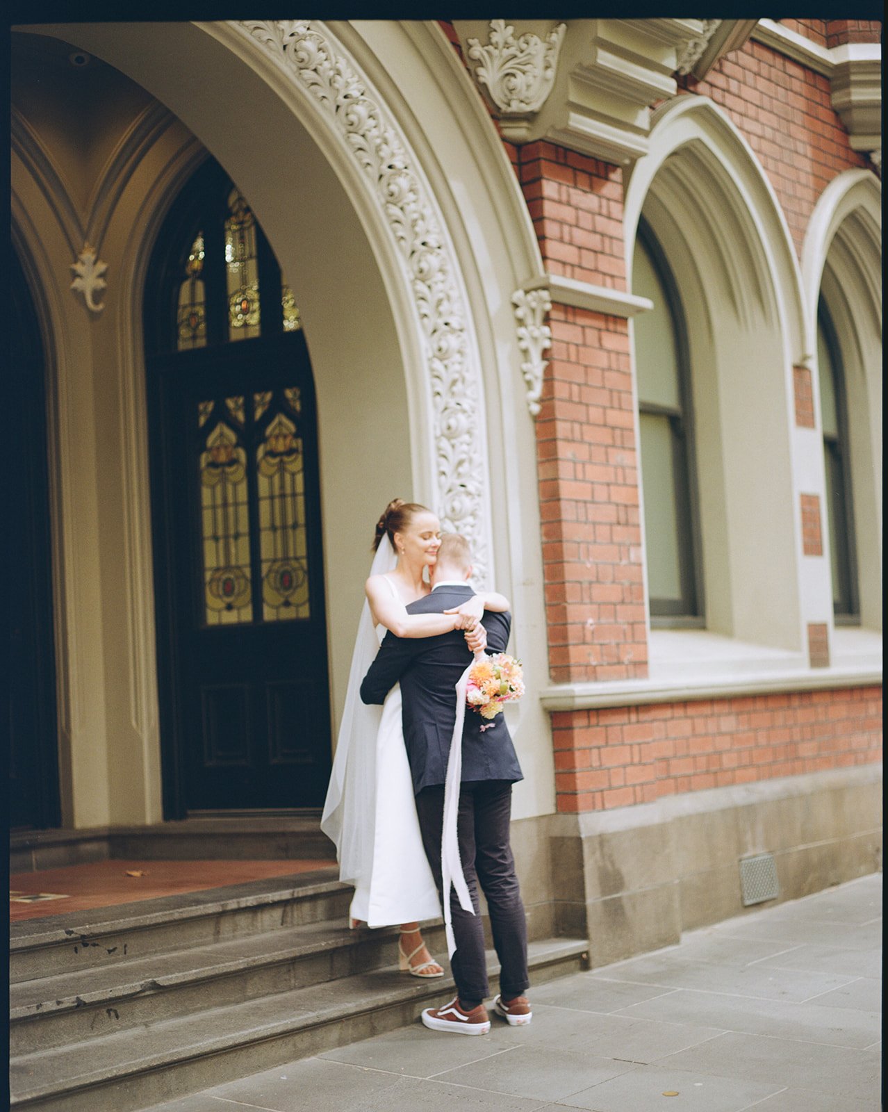 a bride and groom stand on some steps in the city, a glass stained window is behind them. The bride flops her bouquet over the grooms shoulder.