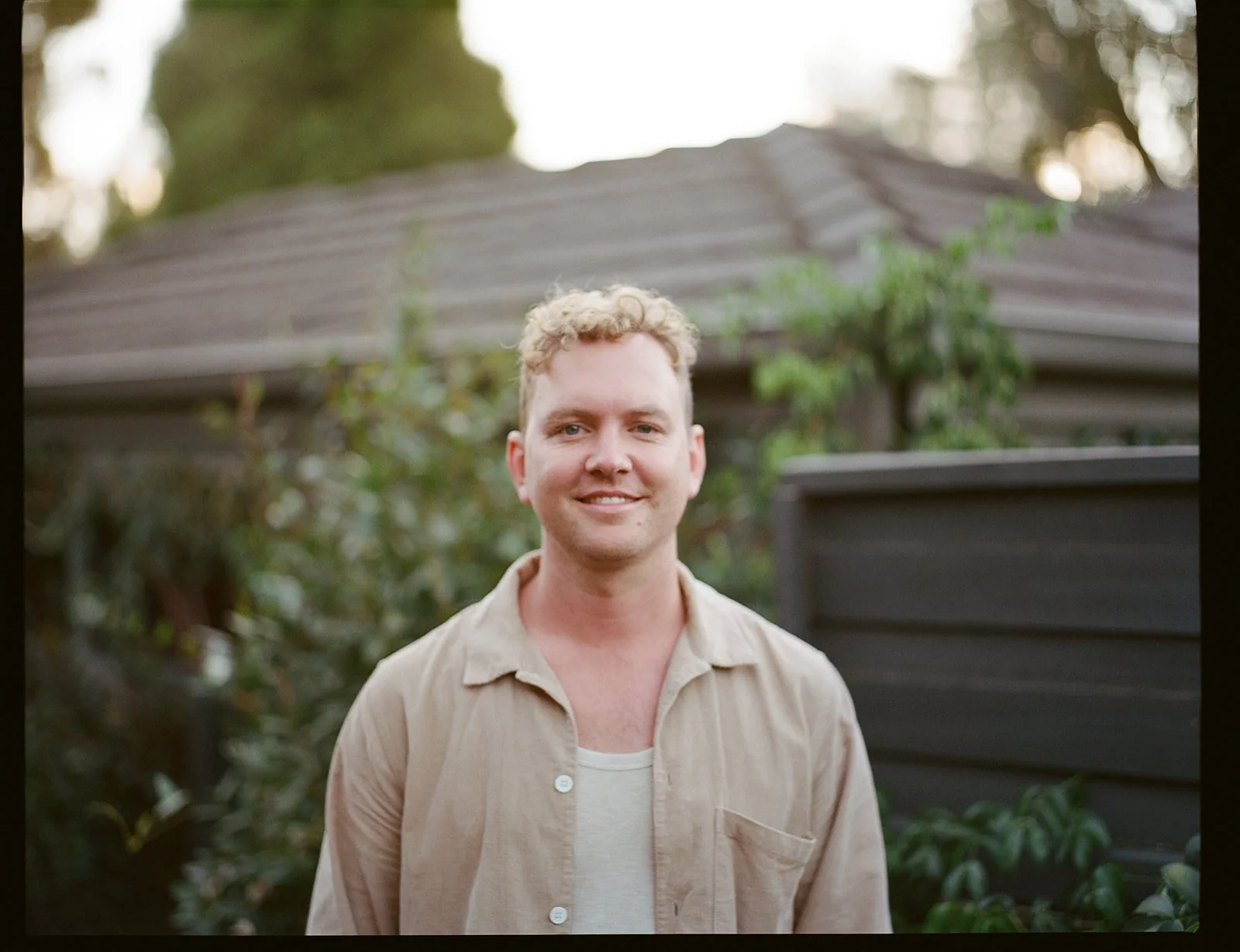 a man stands in a garden in front of a fence, he is wearing a light shirt and smiling directly at the camera.