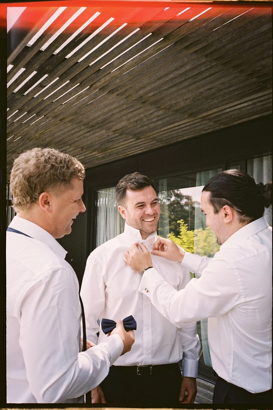 Two groomsmen adjust the grooms top button, they are standing outside and wearing white shirts. 