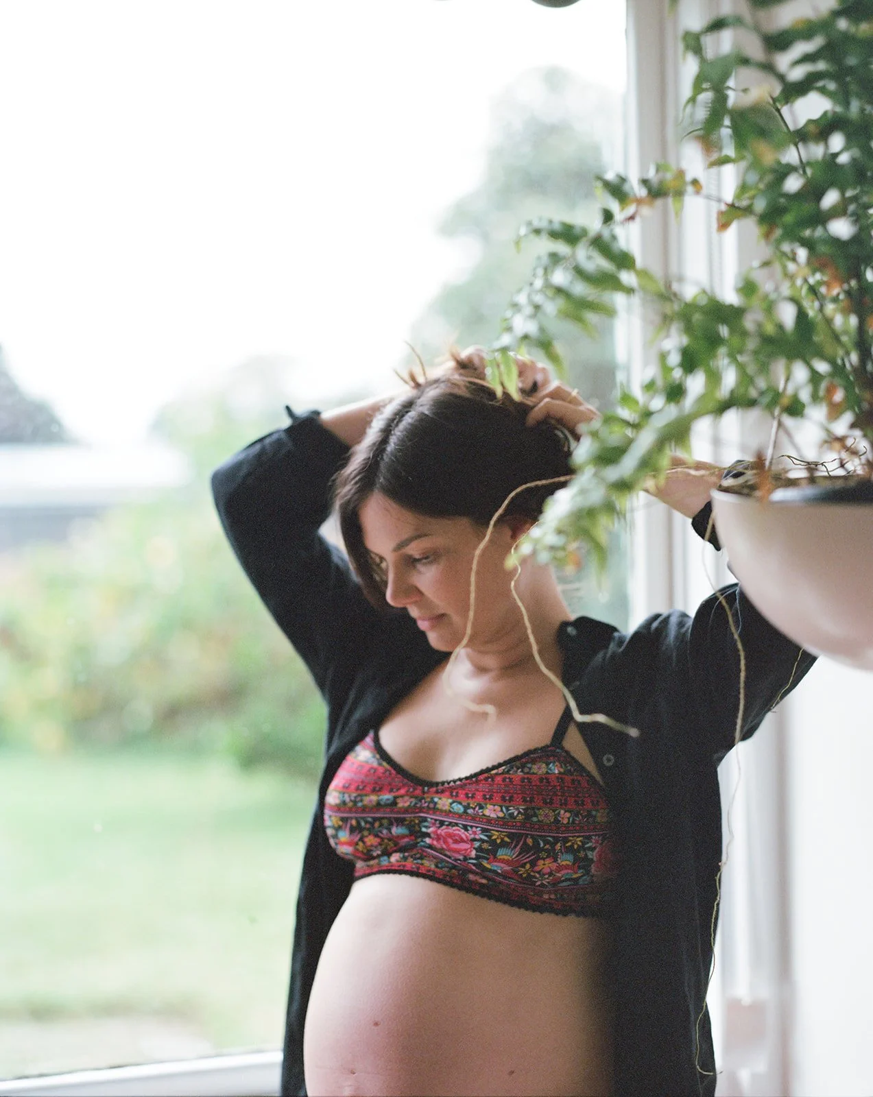 A pregnant lady stand in front of a window, you can see her belly as she looks down and ties up her hair.