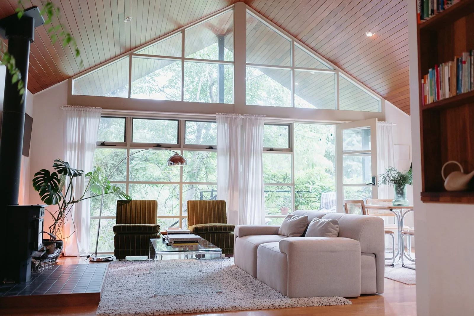 a light and open lounge room with big windows, a cream comfy couch, velvet green arm chairs and a curved silver lamp.