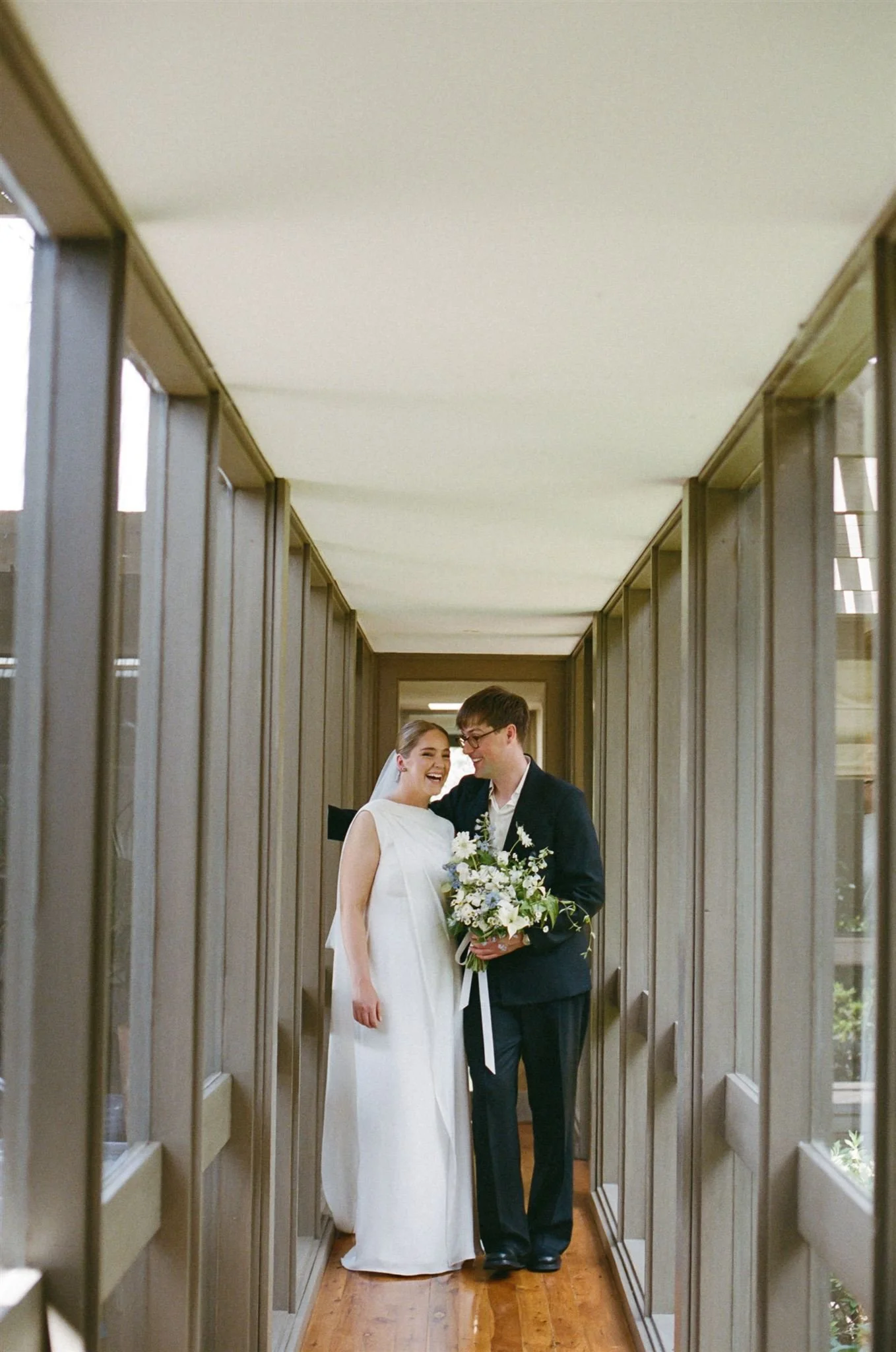 A bride and groom share a happy moment in a narrow hallway, with the bride wearing a white dress and the groom in a dark suit holding a bouquet of flowers.