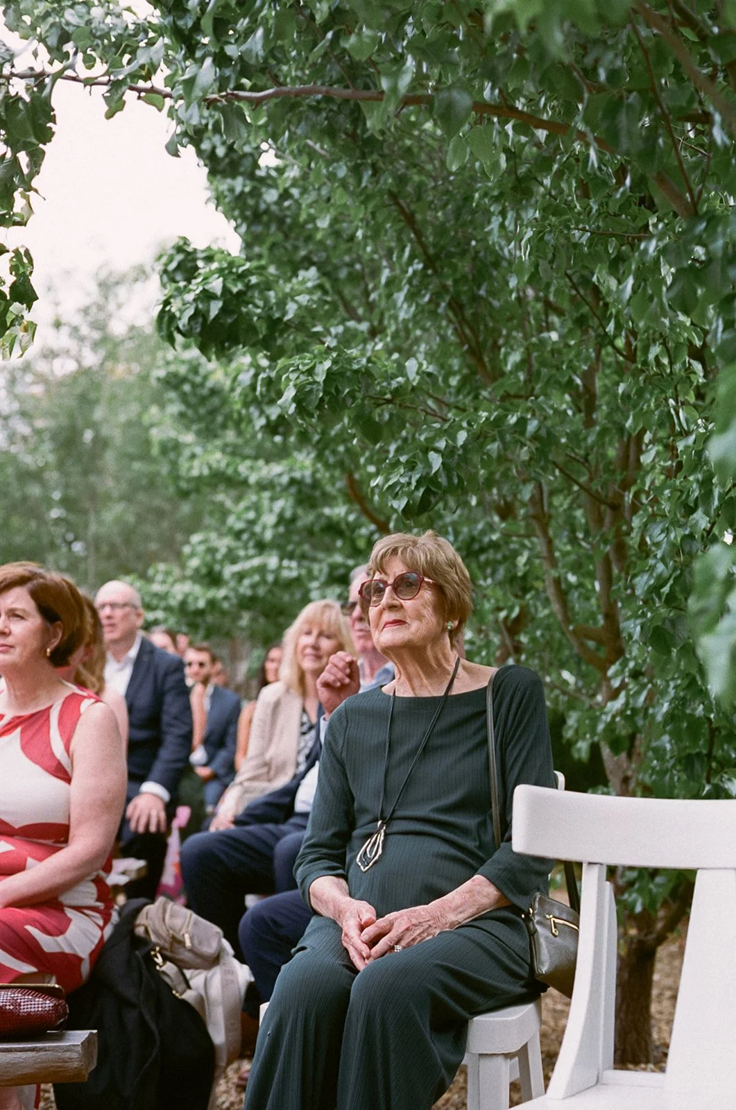 A grandma sits at an outdoor wedding ceremony listening. she is wearing a green top and trousers and sunglasses. 