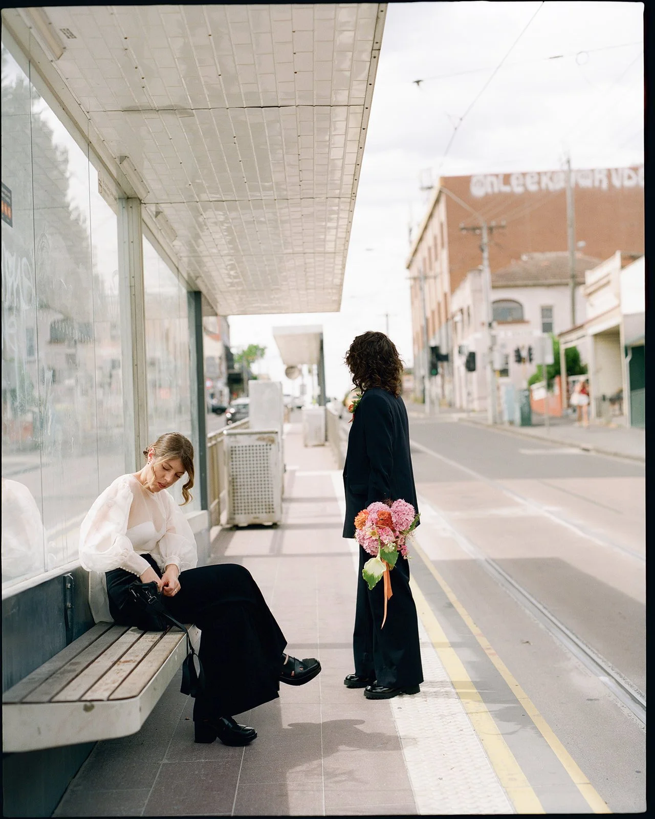 two brides are waiting from a tram in the city, one is sitting down and the other looks down the street holding a pink and orange bouquet.
