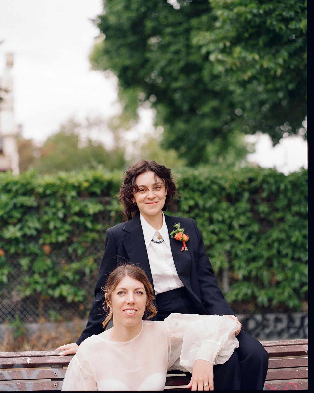 Two brides sit on a bench having their portrait taken. One leans her arm on the others leg. One bride is wearing a black suit and the other is wearing a cream silk blouse. 
