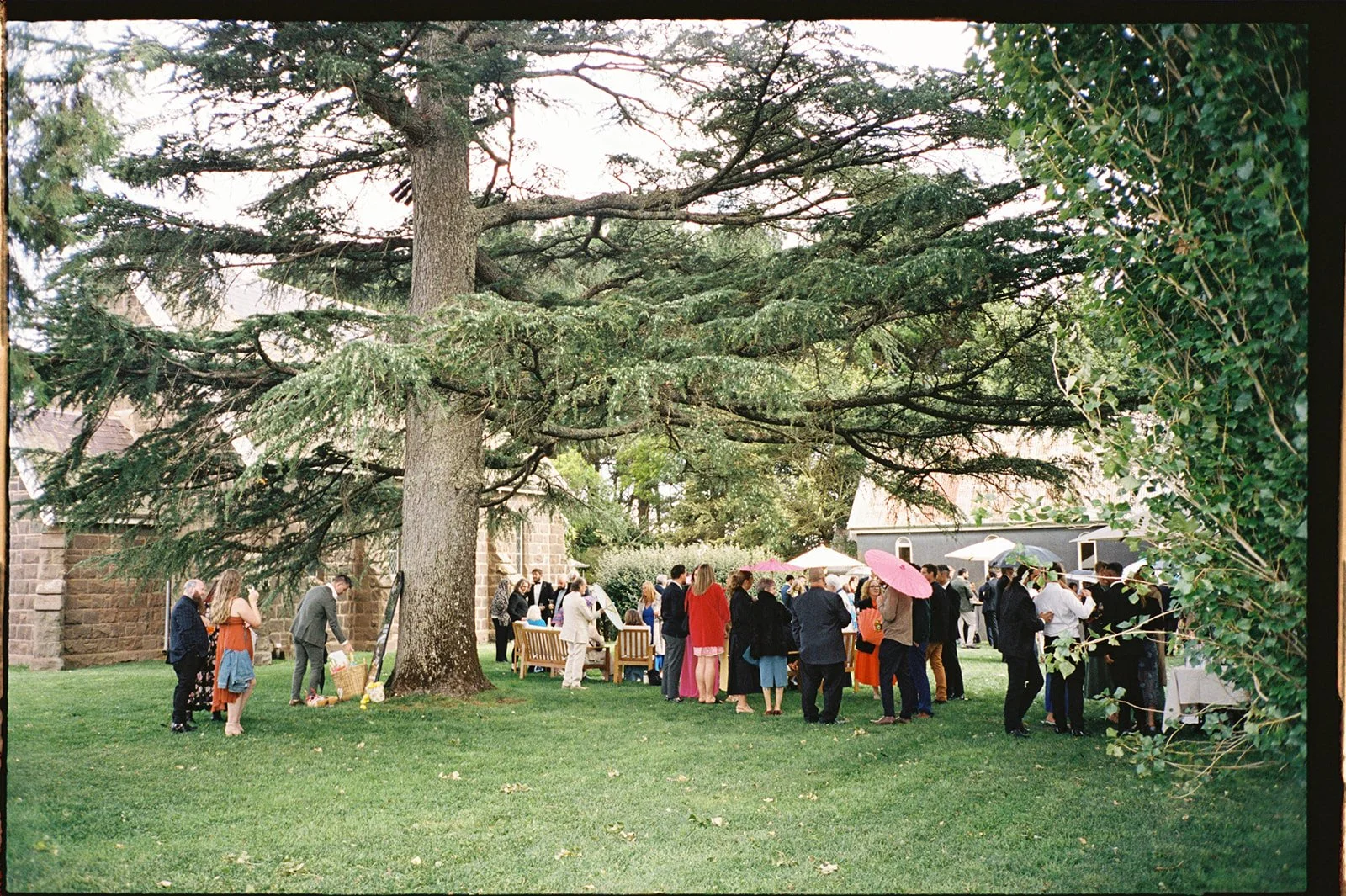 A wedding party gather on the lawn next to a blue stone church, they are wearing colourful outfits.