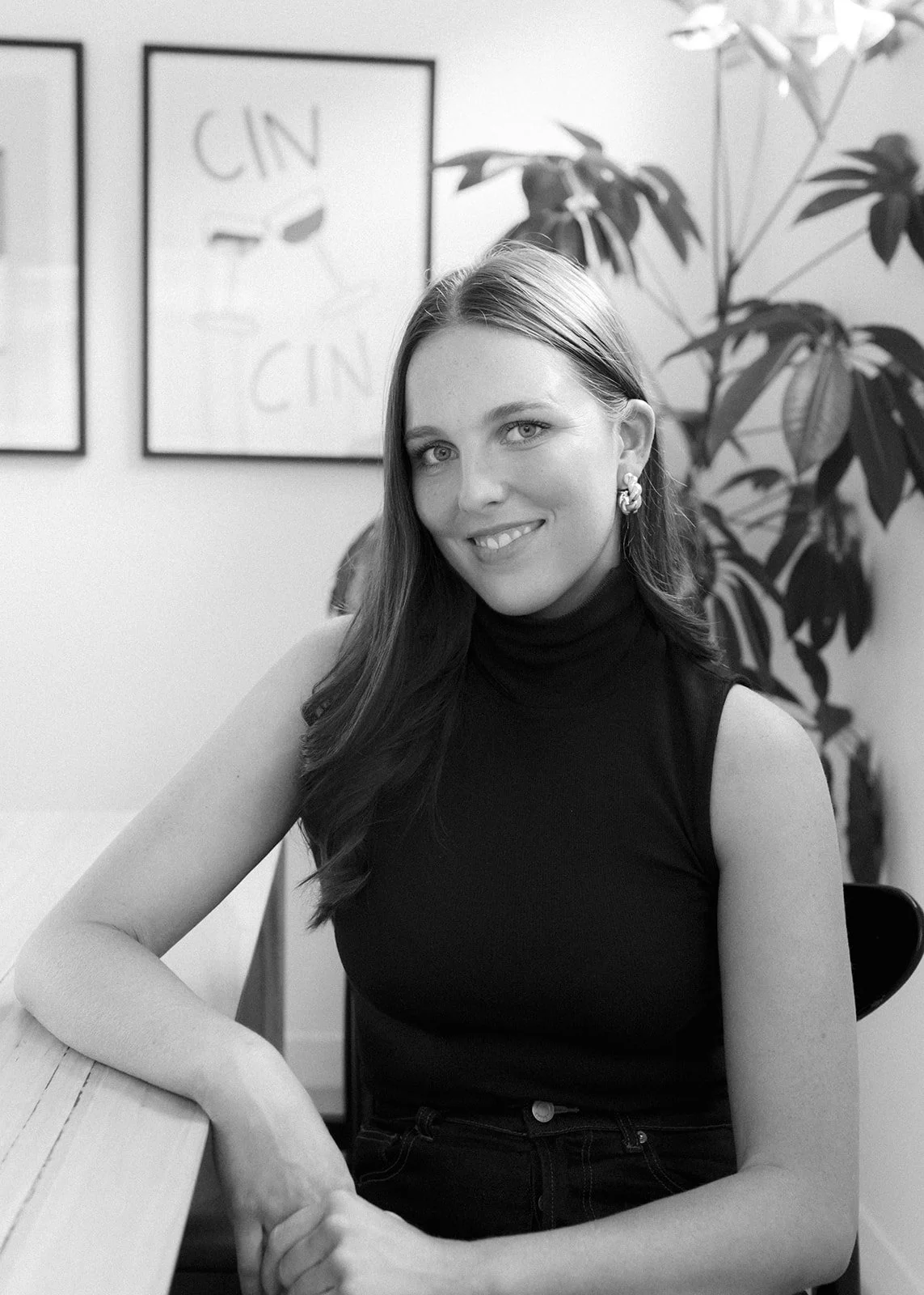a women is sitting and smiling at the camera, she has a black sleeveless top and hoop earrings, the photo is in black and white.