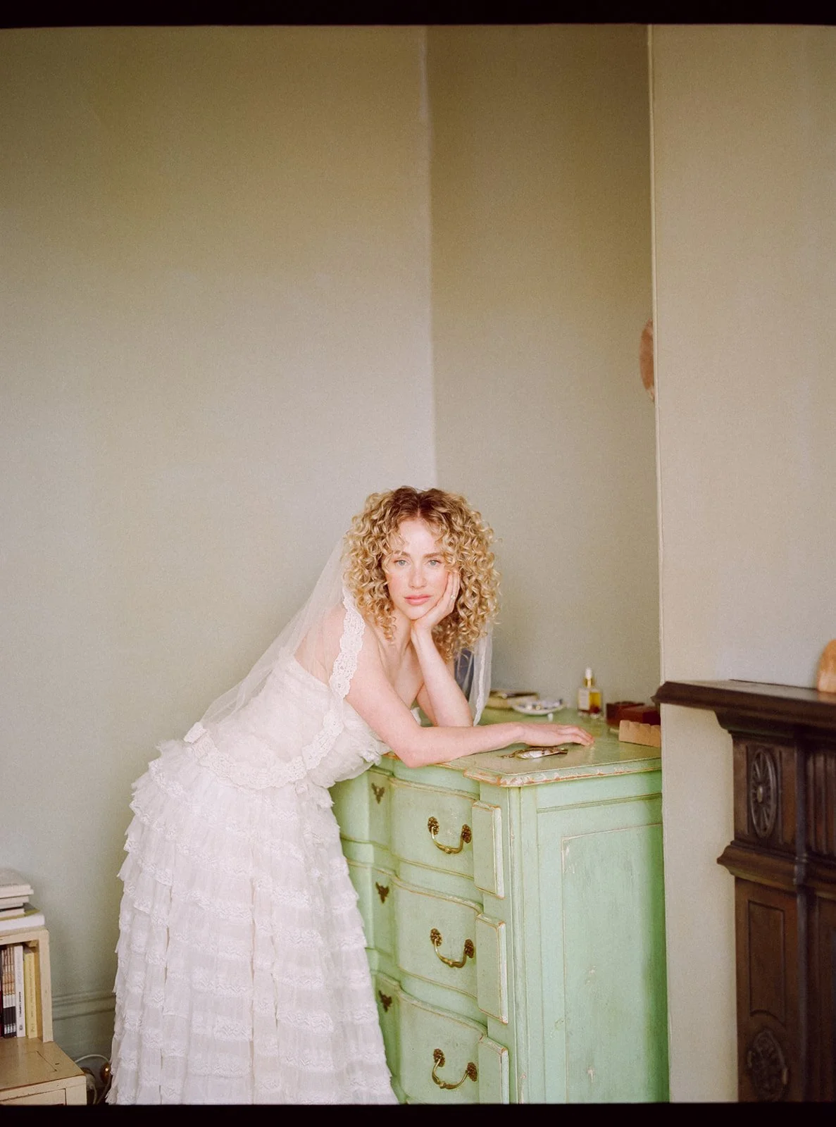 a bride stands in a bedroom against a green chest of drawers, she has curly blonde hair and wears a lace veil.