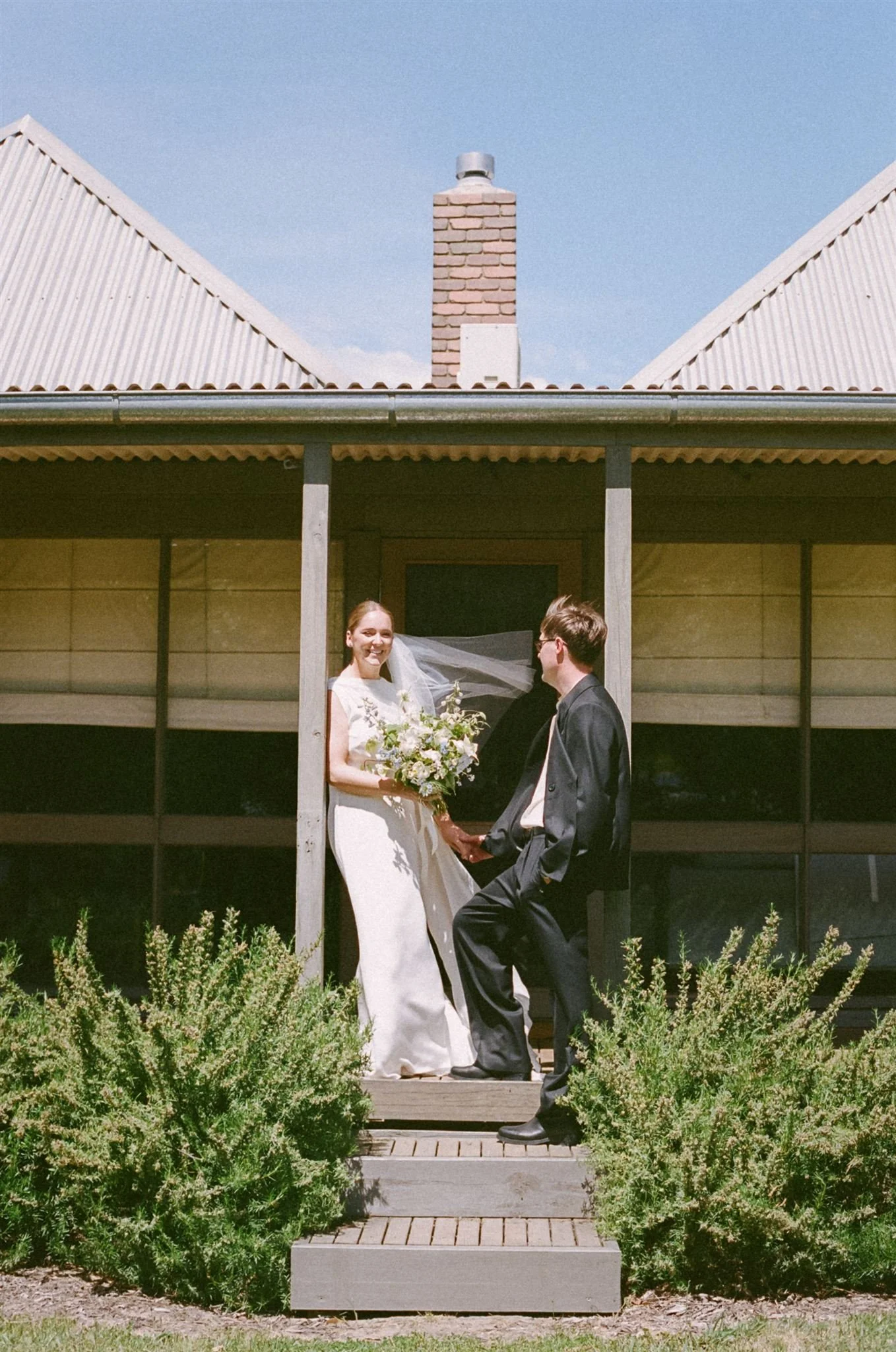 Bride and groom standing on steps outside, the wind is blowing the brides veil, its a sunny day.
