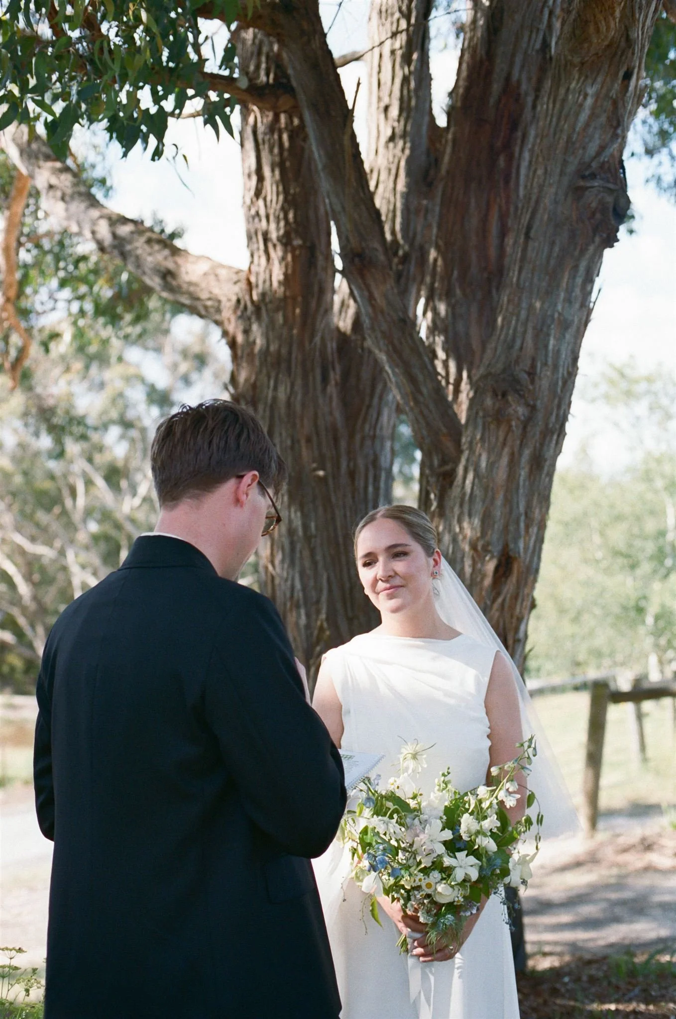 A groom shares his vows with his bride at an outdoor ceremony, she smiles while holding a green, blue, and white bouquet. There is a large tree behind them.