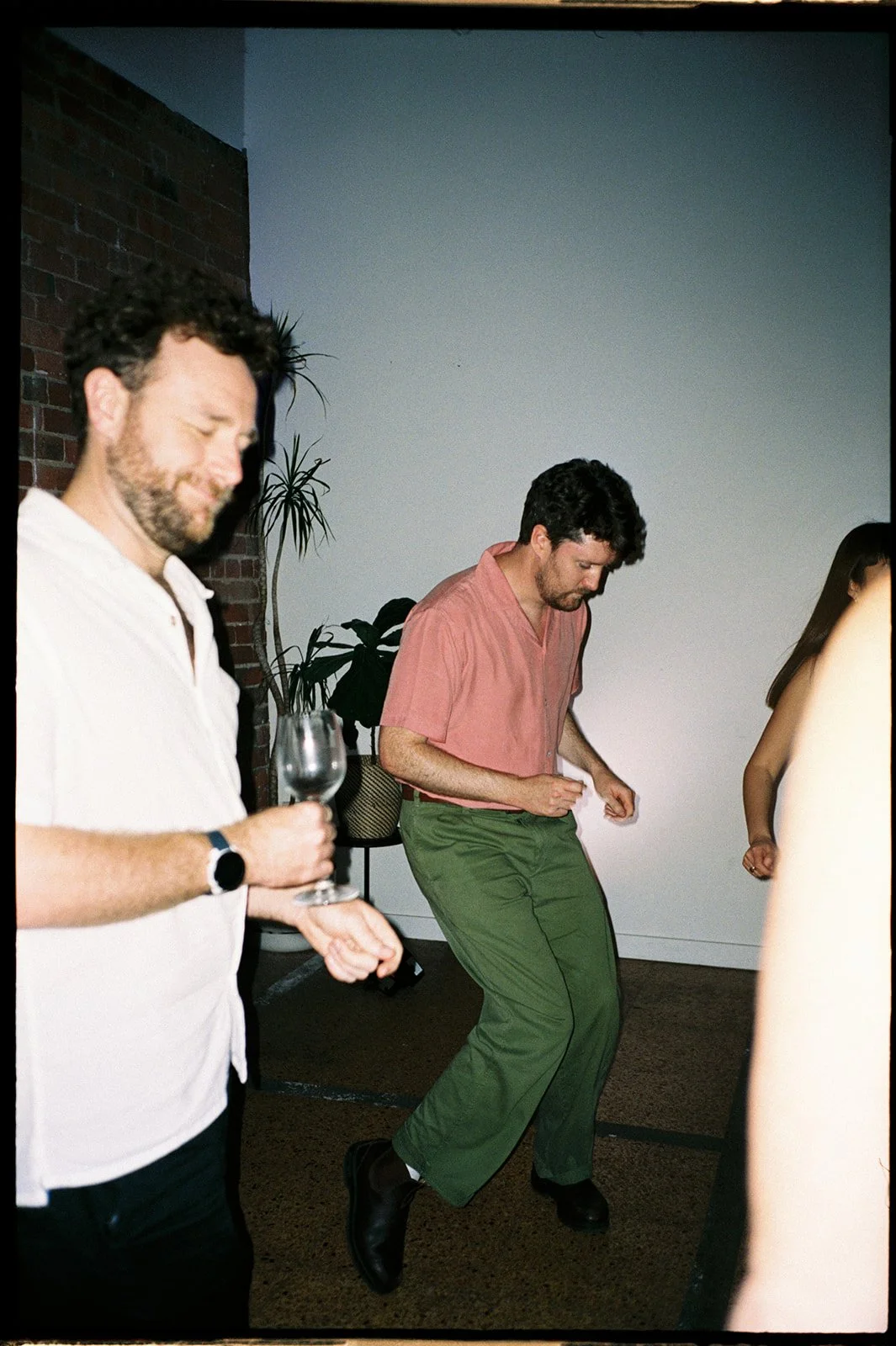 Wedding guests dance at a wedding reception. A guest is wearing a pink shirt and green trousers, the photo is taken with flash.