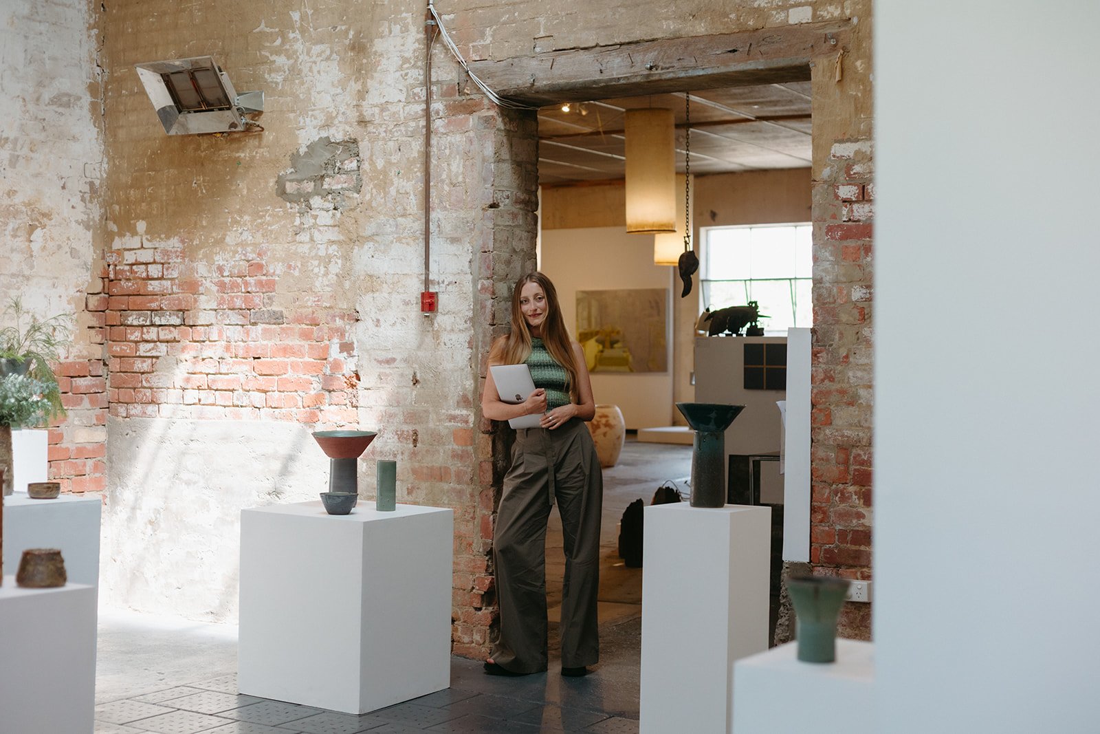 a women stands in an art gallery leaning against a brick wall, she holds a laptop in her arms.