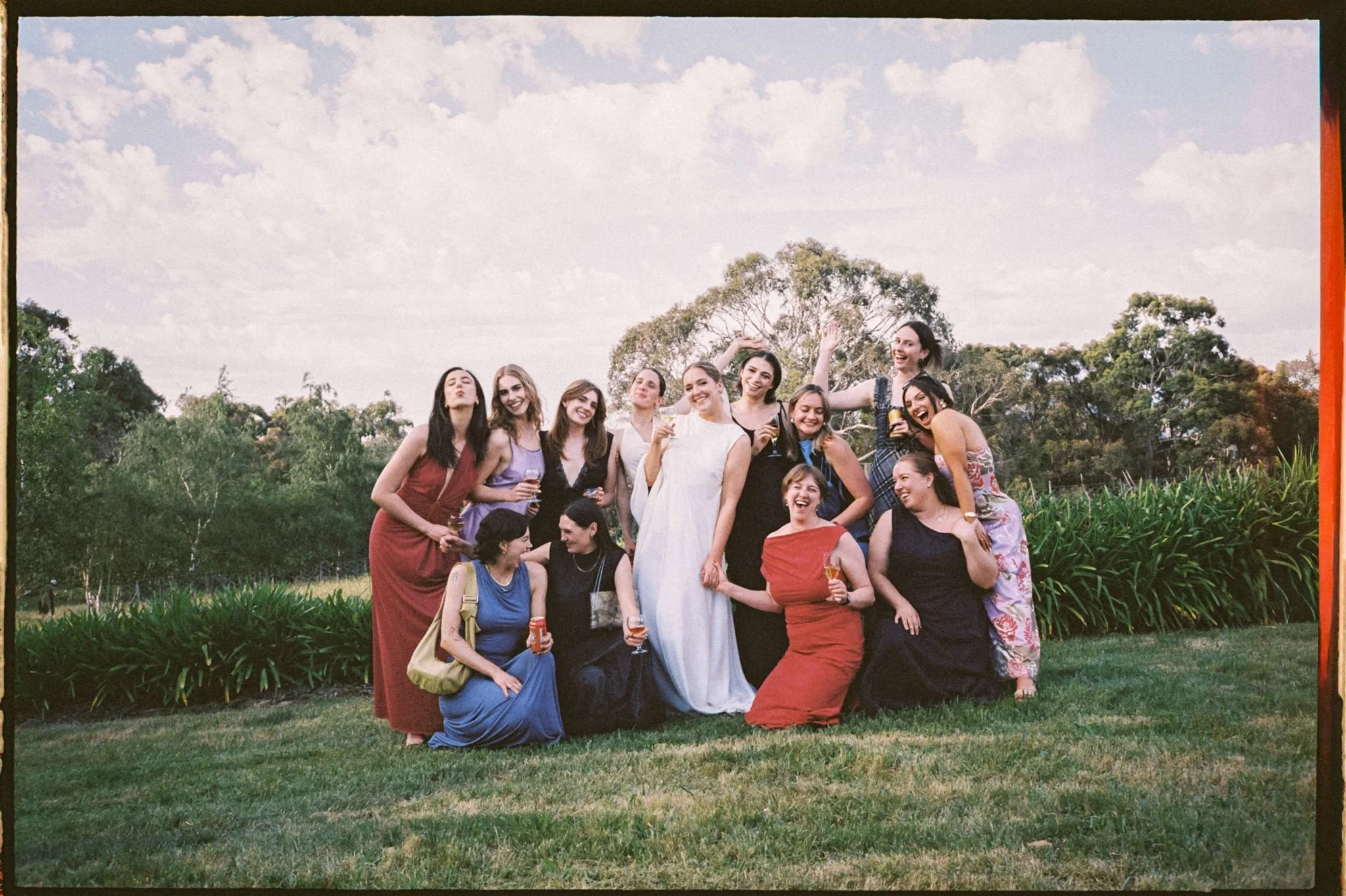 A group of women in colorful dresses outdoors, smiling and holding drinks, gathered for a wedding in a park-like setting with trees and grass.