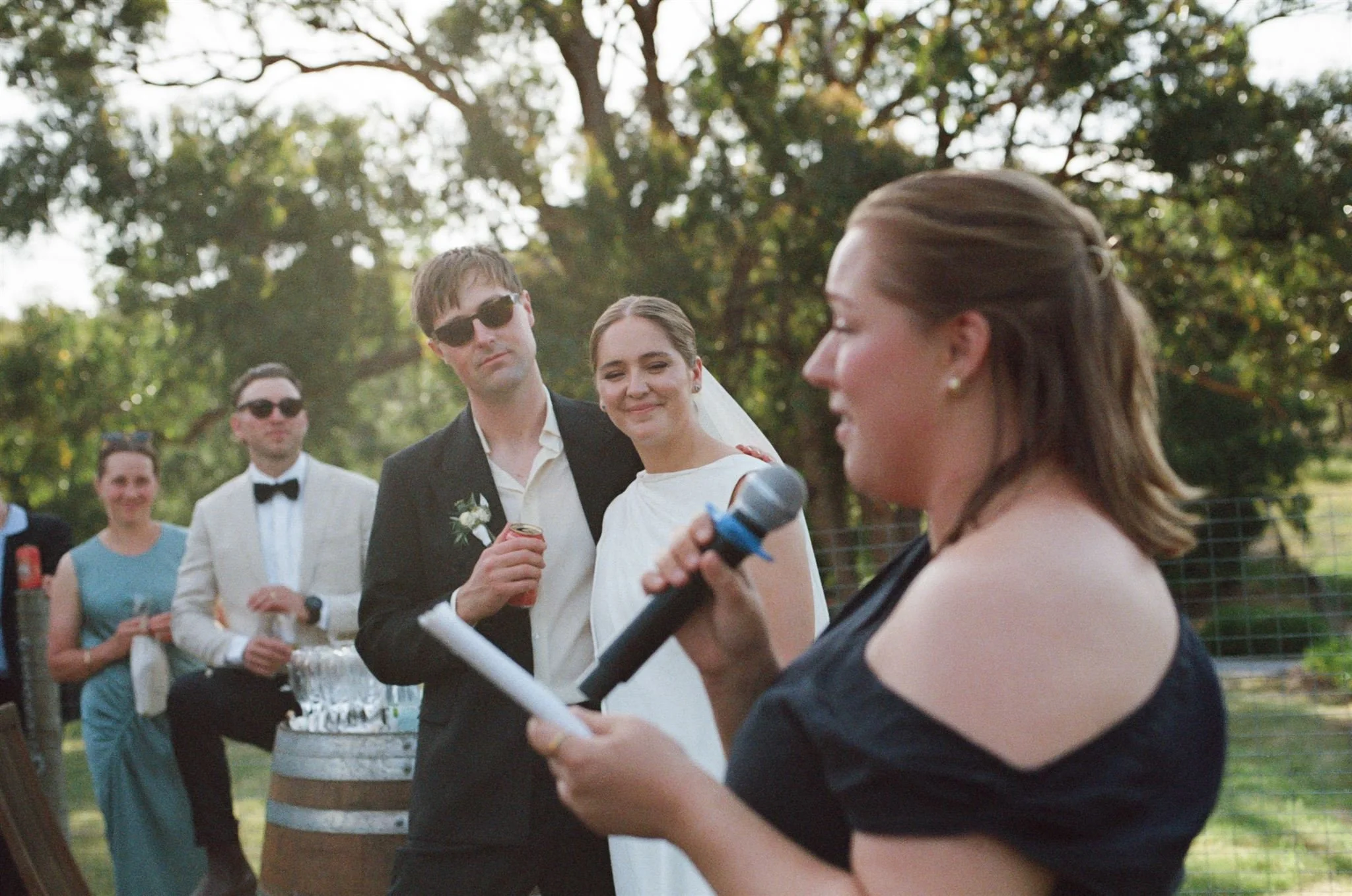 A guest is giving a speech at a wedding while the bride and groom stand in the background smiling. They are standing in a garden. 