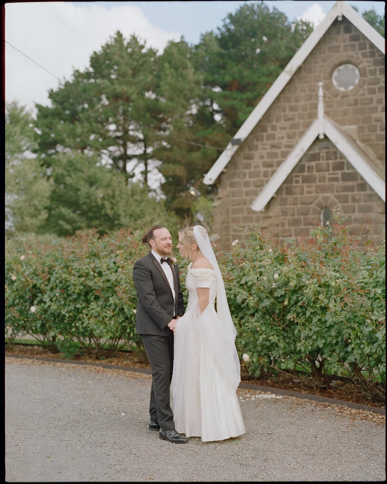 A bride and groom standing outdoors in front of a small stone church with large trees in the background. The groom is wearing a black tuxedo with a bow tie, and the bride is wearing a white wedding dress with puffed sleeves. The sky is partly cloudy.