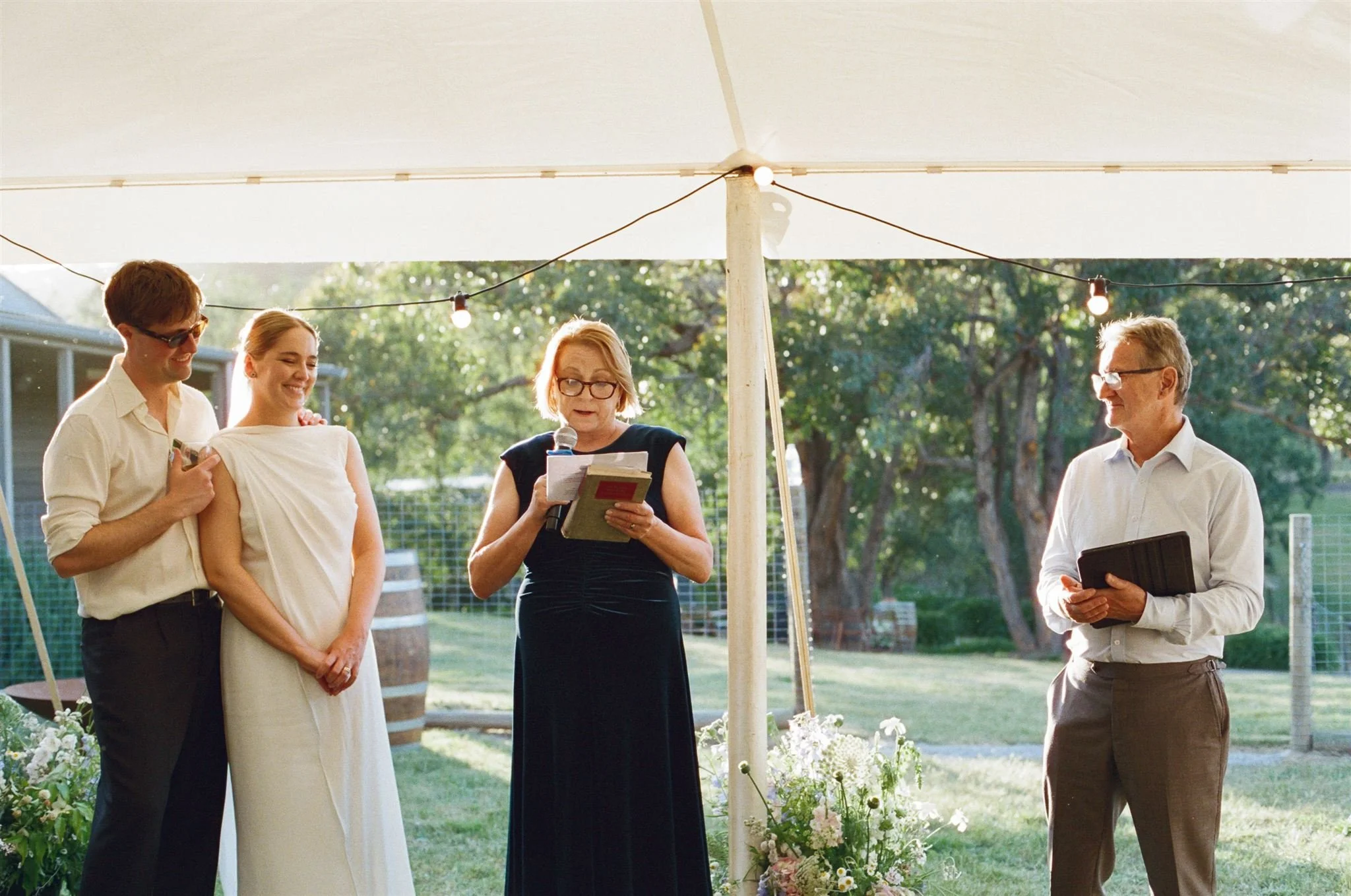A bride and groom standing next to the brides parents while they give their wedding speech. Its golden hour and they all have a warm glow.