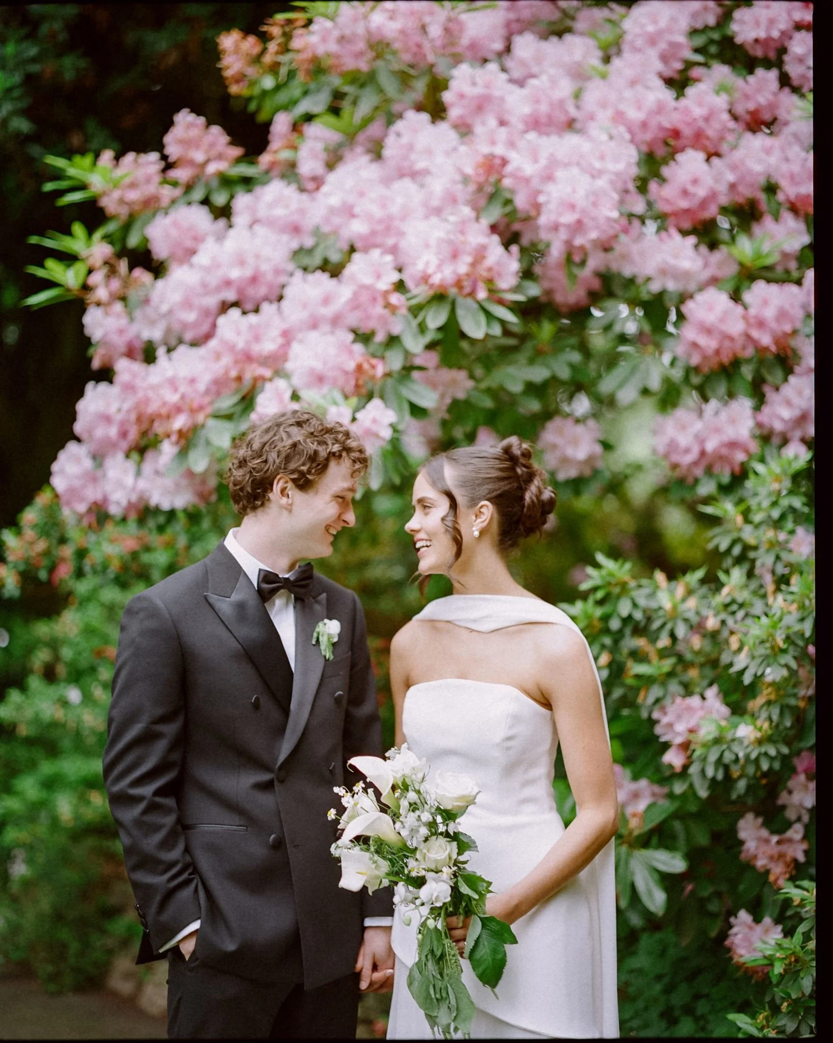 A bride and groom stand in front of a bush with pink flowers. They are smiling at each other, the grooms wears a black double breasted jacket and the bride wears a sleeveless white dress with silk scarf.