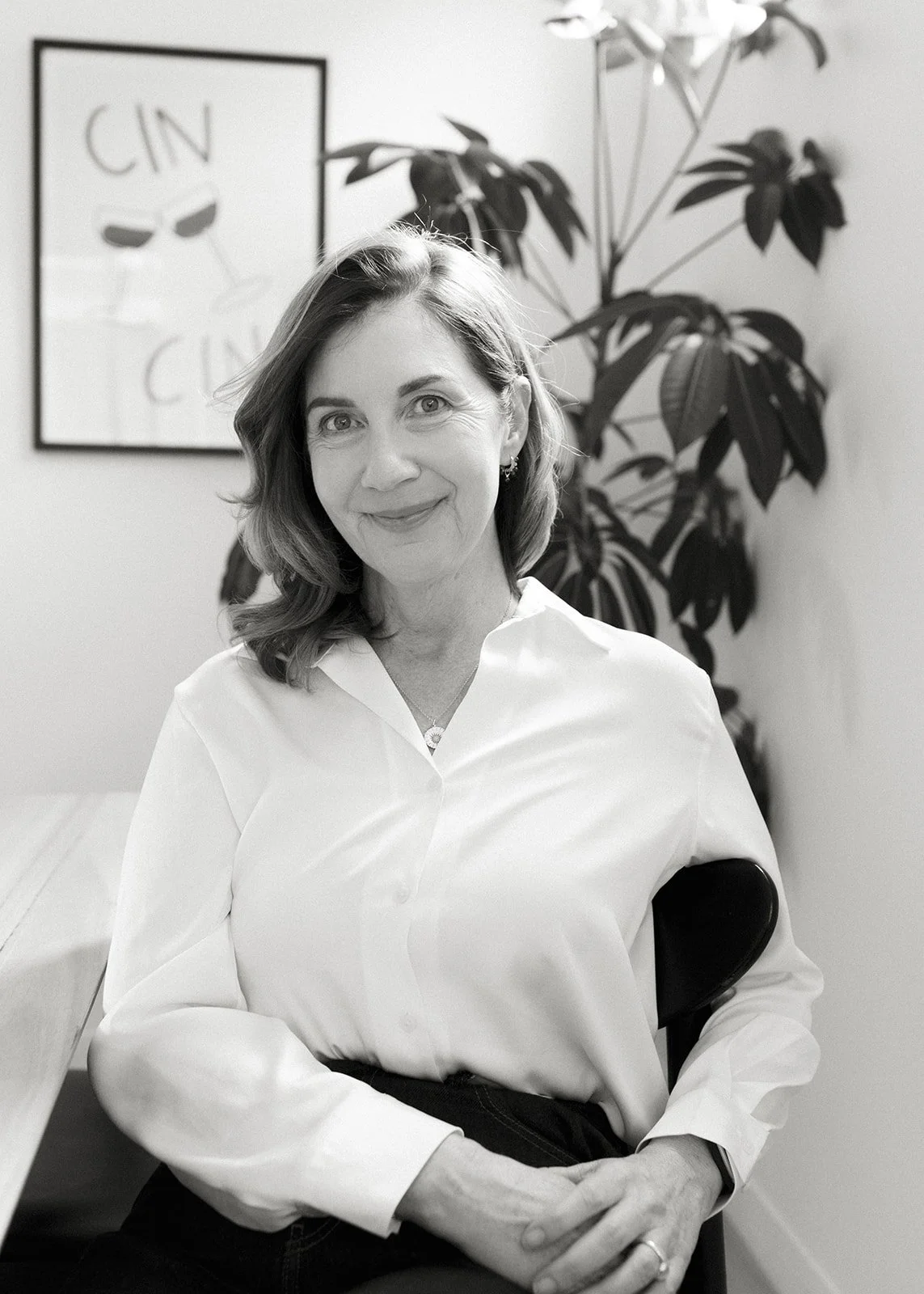 a women sits smiling at the camera, she wears a white shirt and there is a plant in the background, the photo is in black and white.