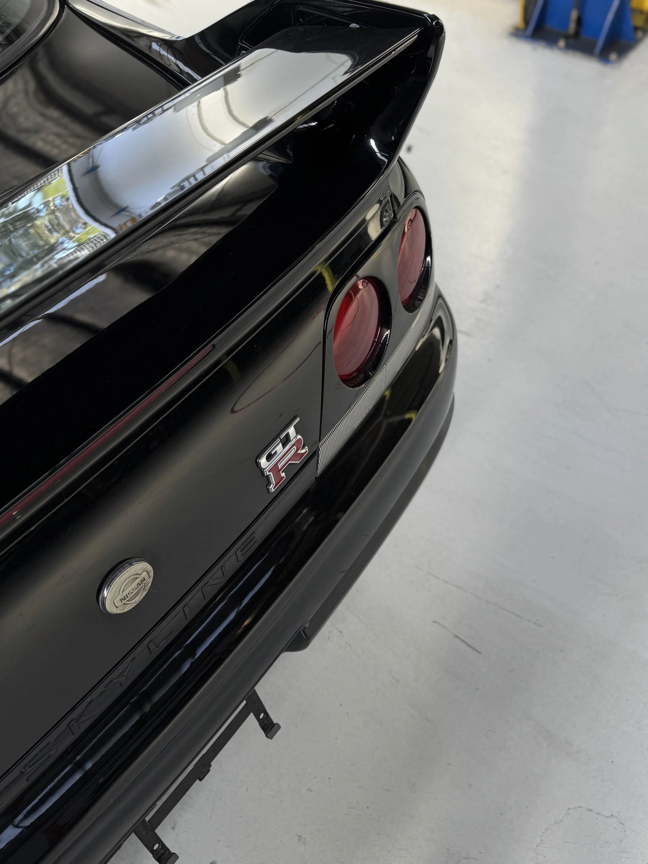 Close-up of the rear of a black Nissan GT-R car, showing taillights and Nissan badge, in a garage or workshop setting.