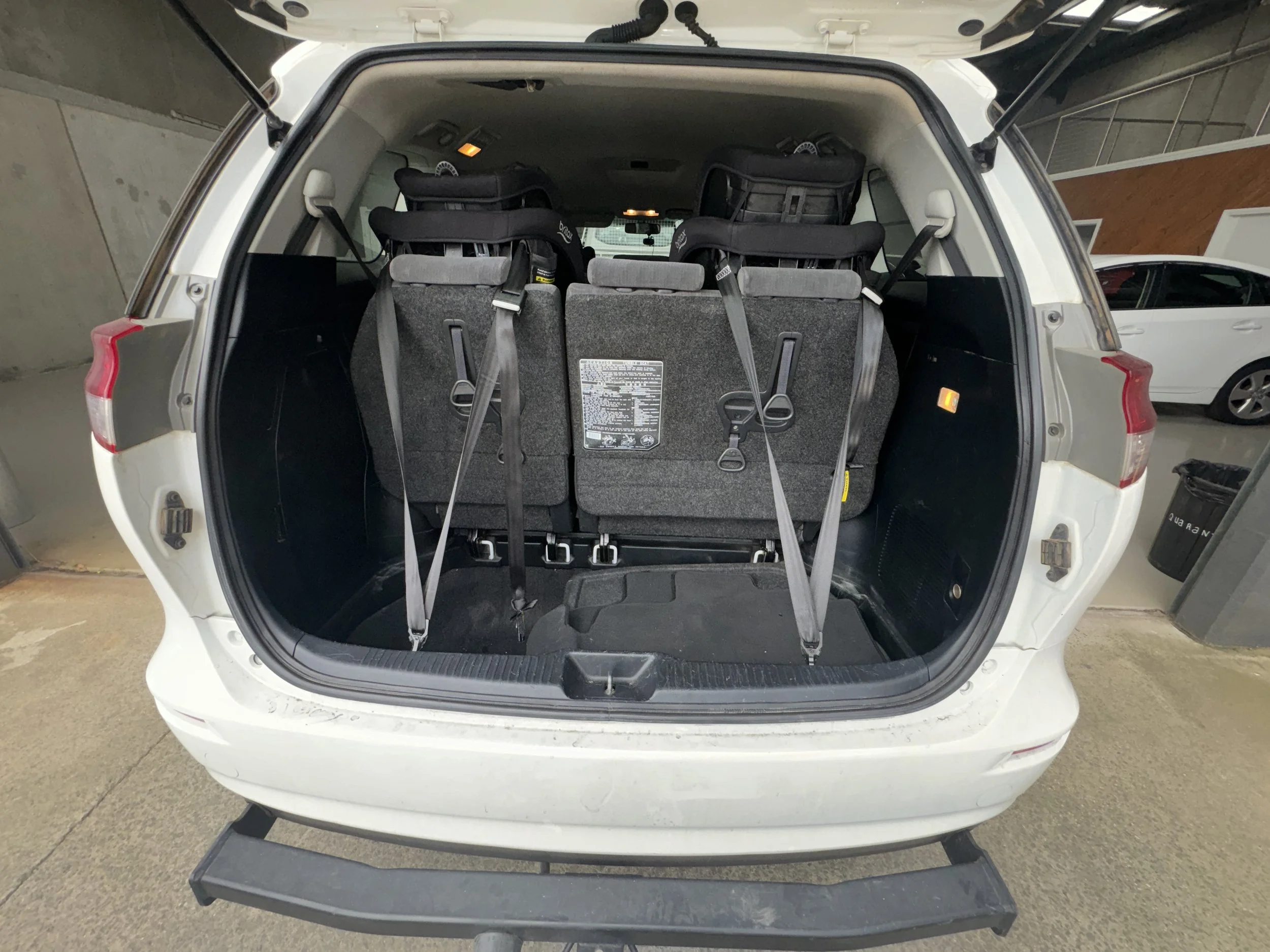 The open trunk of a white SUV showing the back seats folded up and secured, with three car seats installed on the last row, child restraints installed, engineered and certified by VASS1089 with a VASS certificate and modification plate.