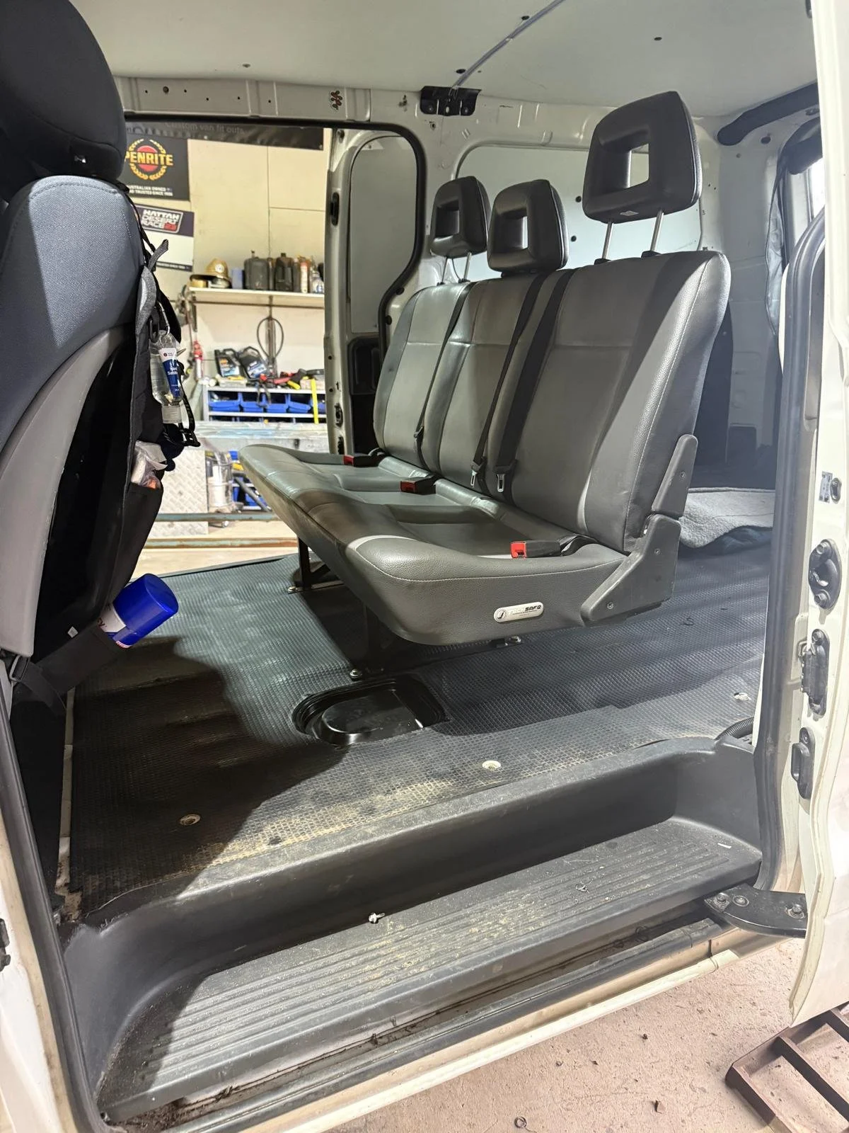 Interior of a vehicle showing gray seats with seat belts, black floor, and a workshop in the background.