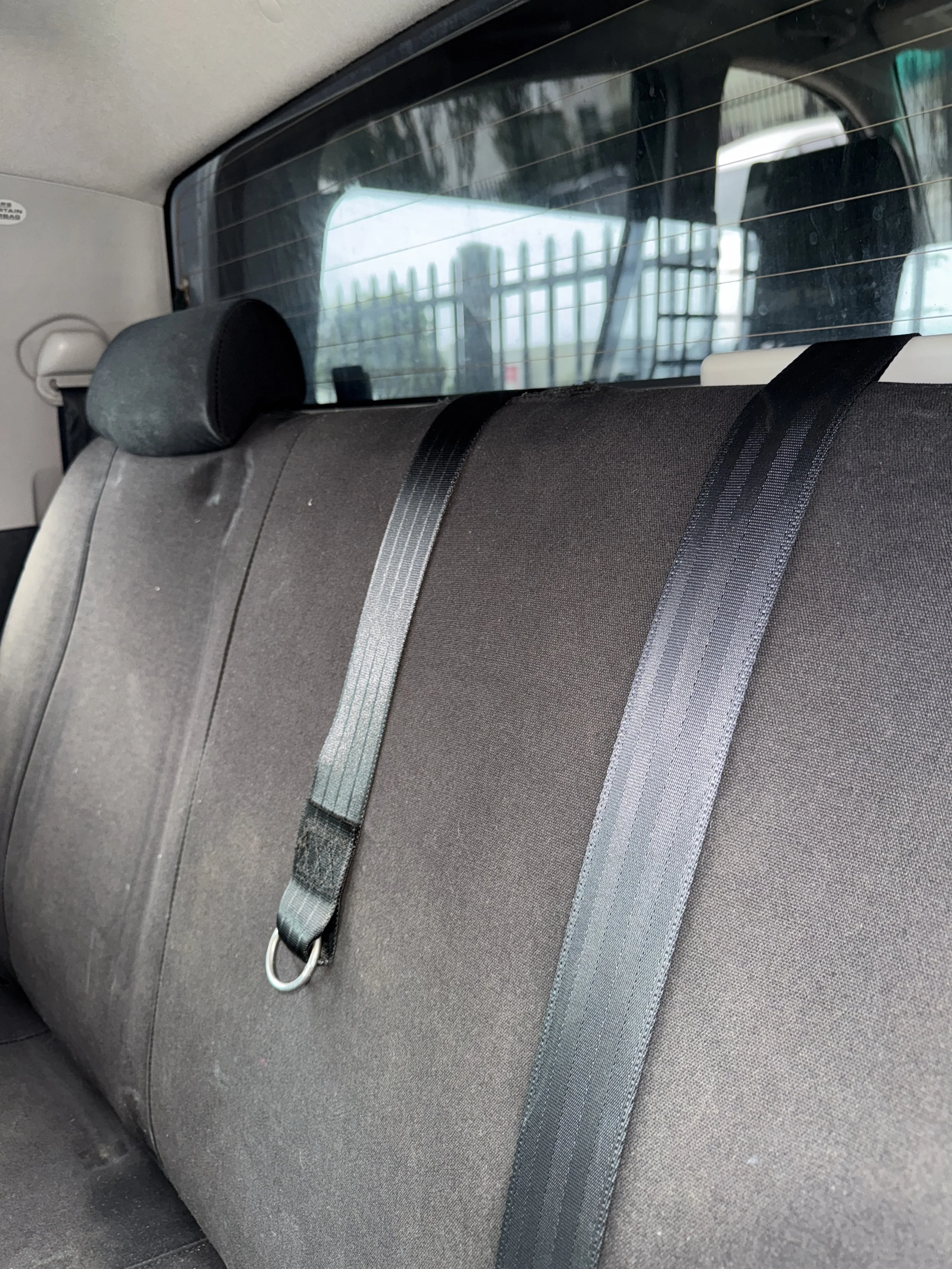 Back seat of a vehicle with a headrest and seat belts, viewed through the rear window with a fence and outdoor area visible outside.