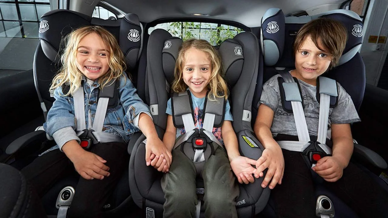 Three children sitting in car seats inside a vehicle, holding hands, with the two girls smiling and the boy looking at the camera.