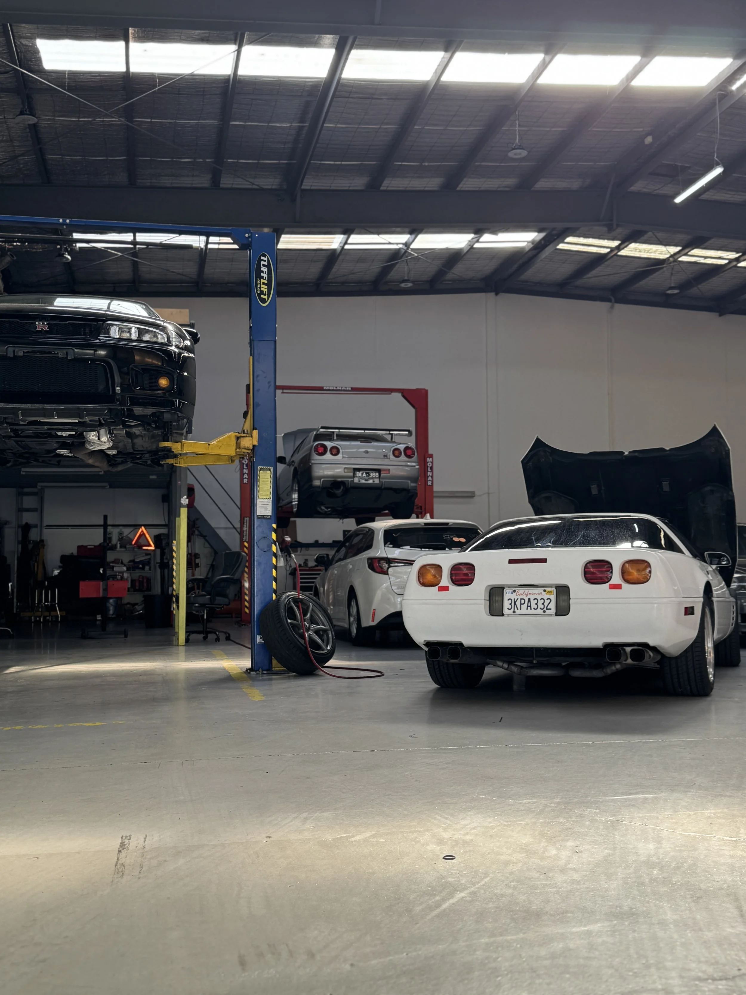 A garage with a black R33 GTRcar on a hydraulic lift, a silver sports car parked below the lift, and a white car in the foreground, with a car hood on the right side.