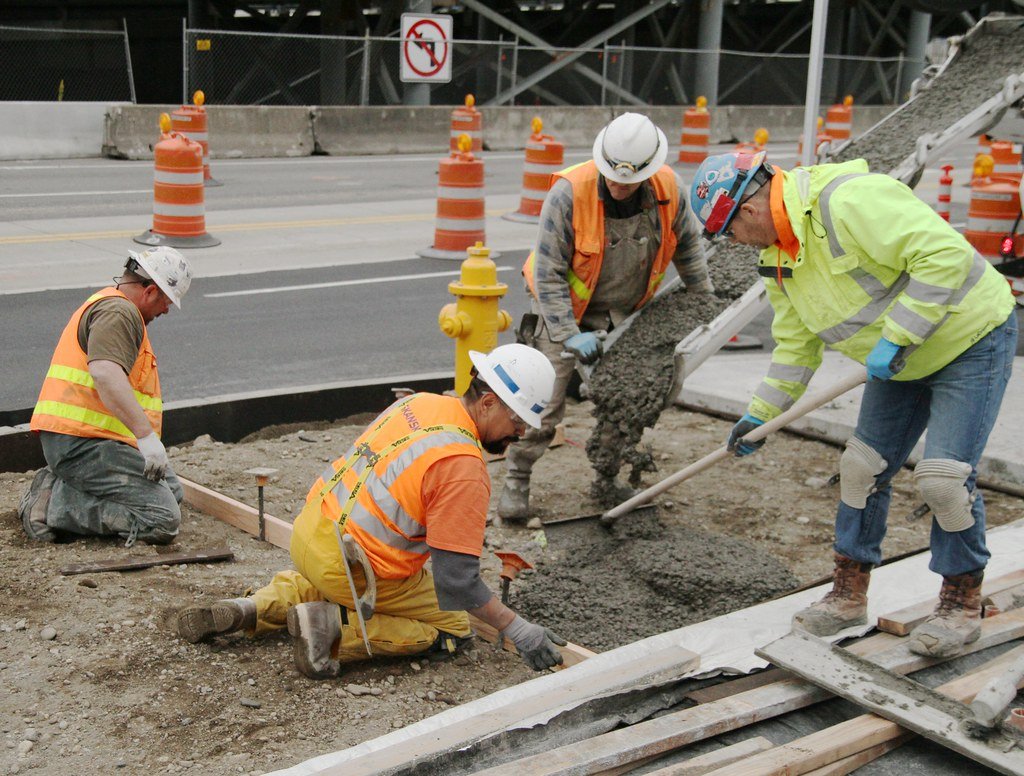 Construction workers pouring concrete on a road site wearing safety gear and helmets.