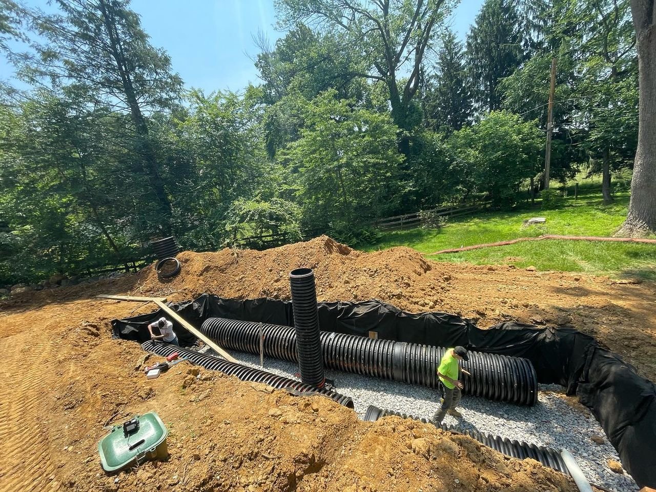 Excavation site with large black pipes being installed in the ground, surrounded by dirt and trees. A person is working nearby.