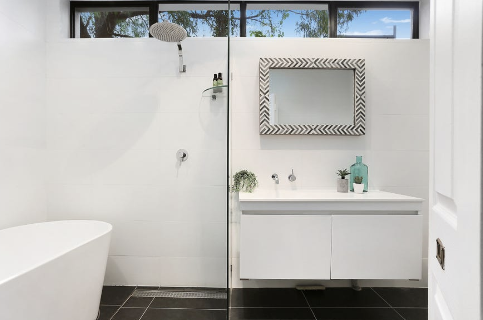 Modern bathroom with a walk-in shower area featuring a rainfall showerhead, a bathtubs, a white vanity with a mirror, and decorative plants, with natural light coming through a window at the top.