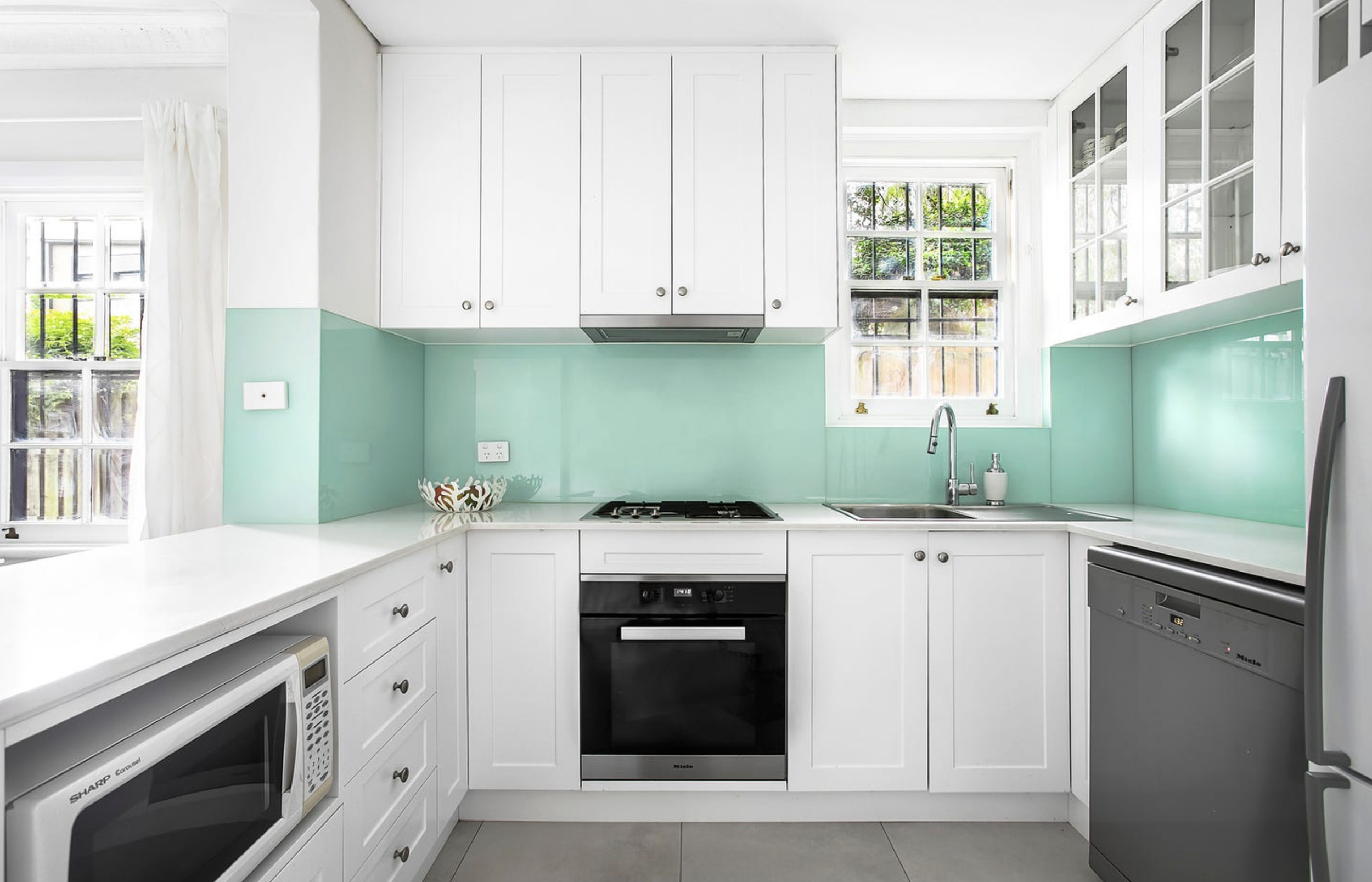 White kitchen with mint green backsplash, white cabinets, black oven, microwave, dishwasher, and a sink near a window with green trees outside