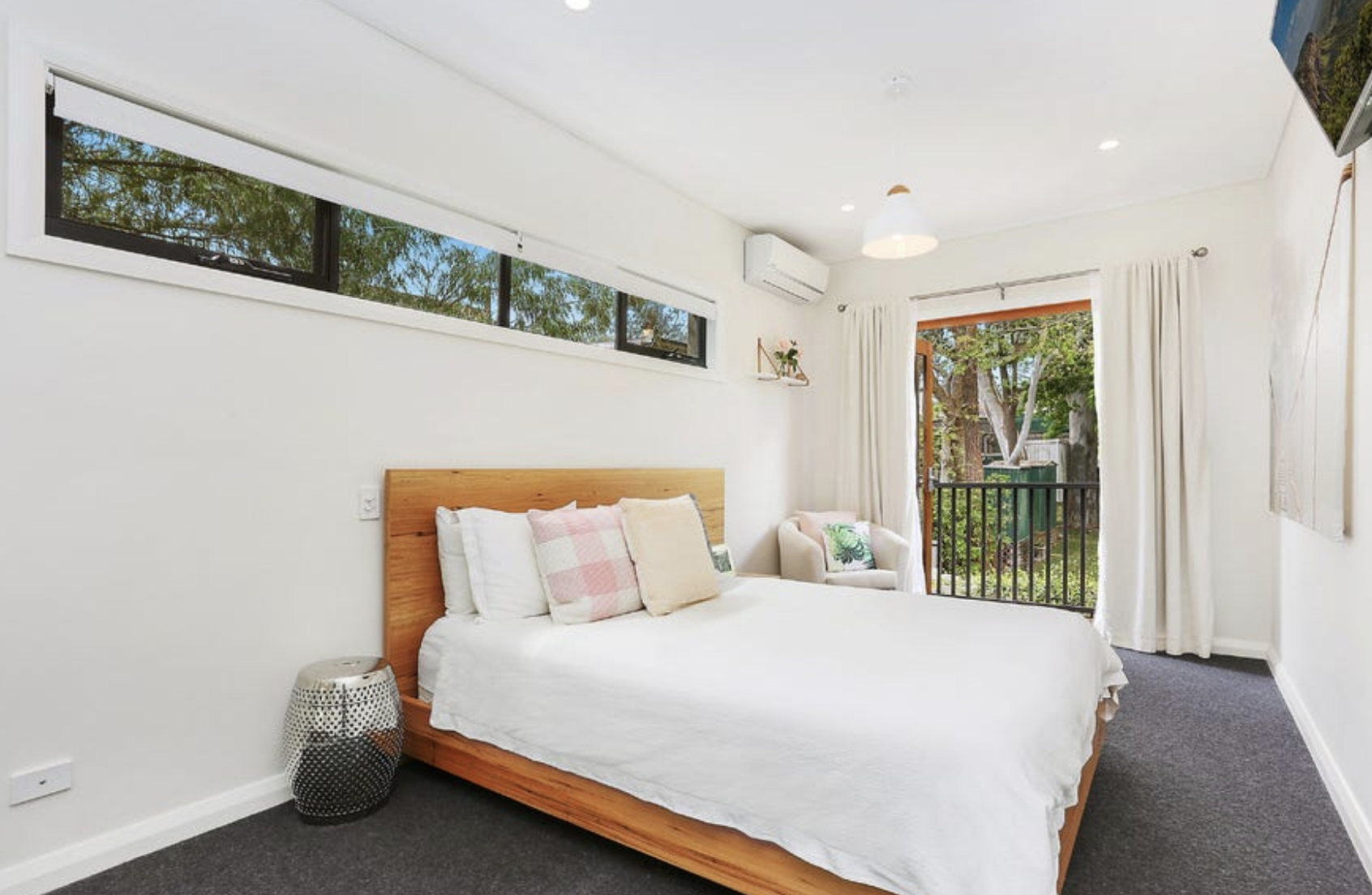 Bright bedroom with a wooden bed frame, white bedding, pink and beige pillows, a small side table, and a sitting armchair near a sliding glass door opening to a view of green trees.