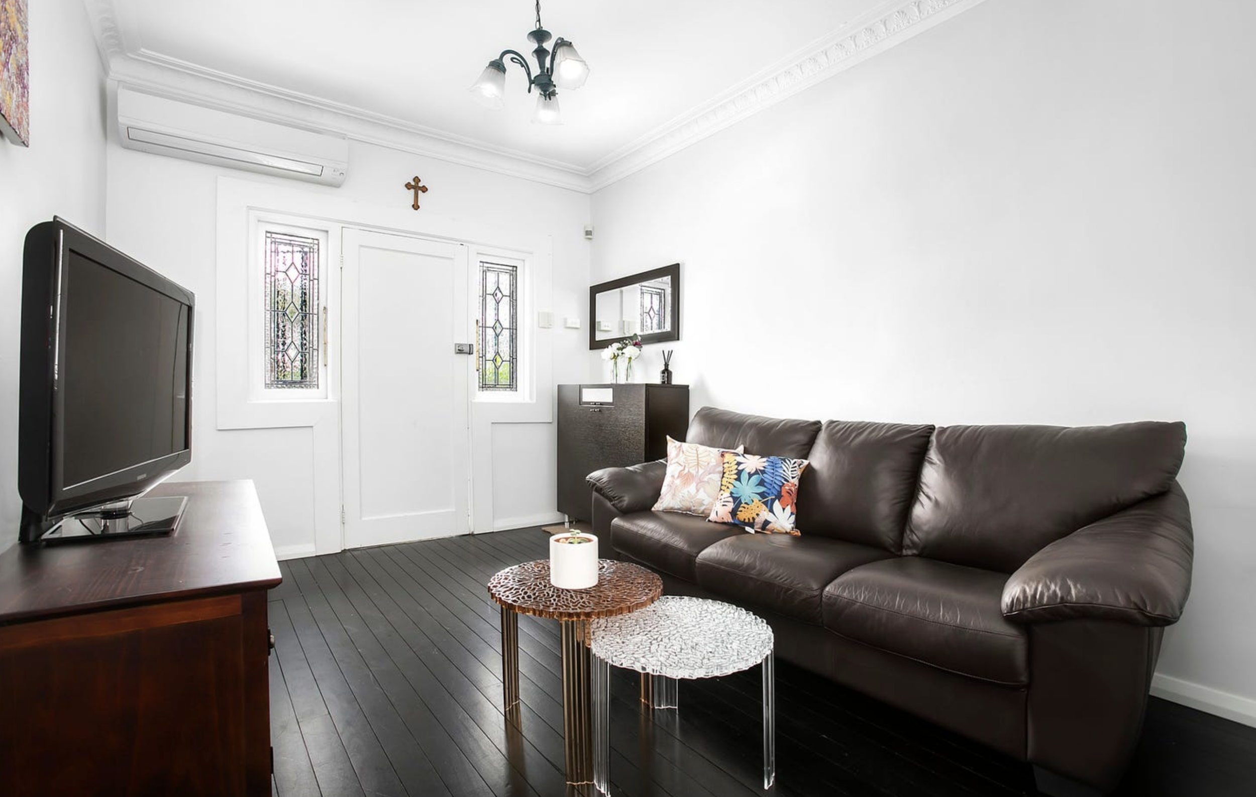Living room with white walls, dark wood flooring, a brown leather sofa with colorful pillows, a small round coffee table, a black cabinet, and a TV on a wooden stand. There are stained glass windows, a mirror, a small vase with flowers, a wall cross, and an air conditioner.